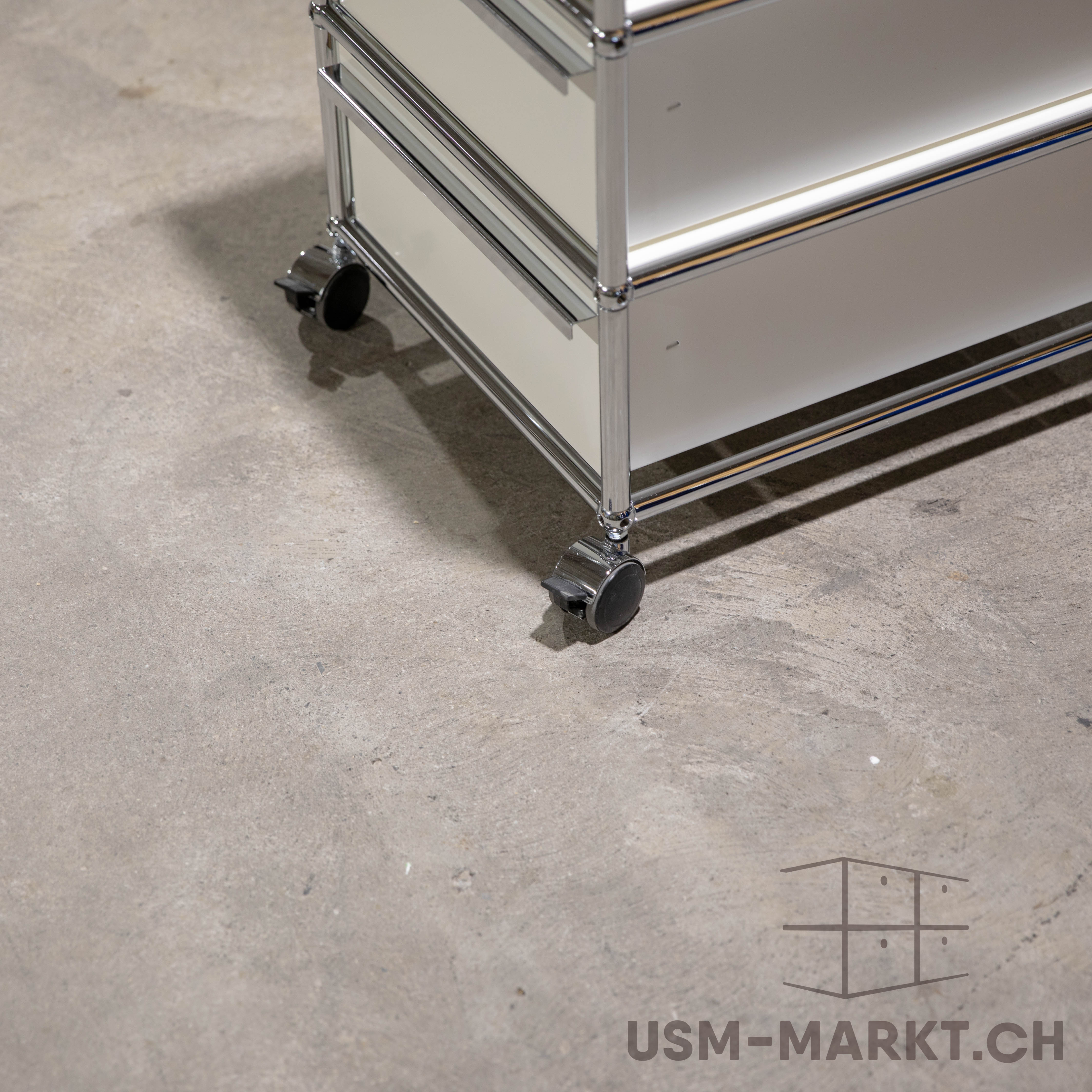 Vista dettagliata di un moderno mobile USM Haller a due livelli su rotelle, di colore chiaro e con telaio cromato lucido. È su un pavimento in cemento grigio chiaro con lievi irregolarità e una linea d'ombra definita; visibili texture e riflessi.