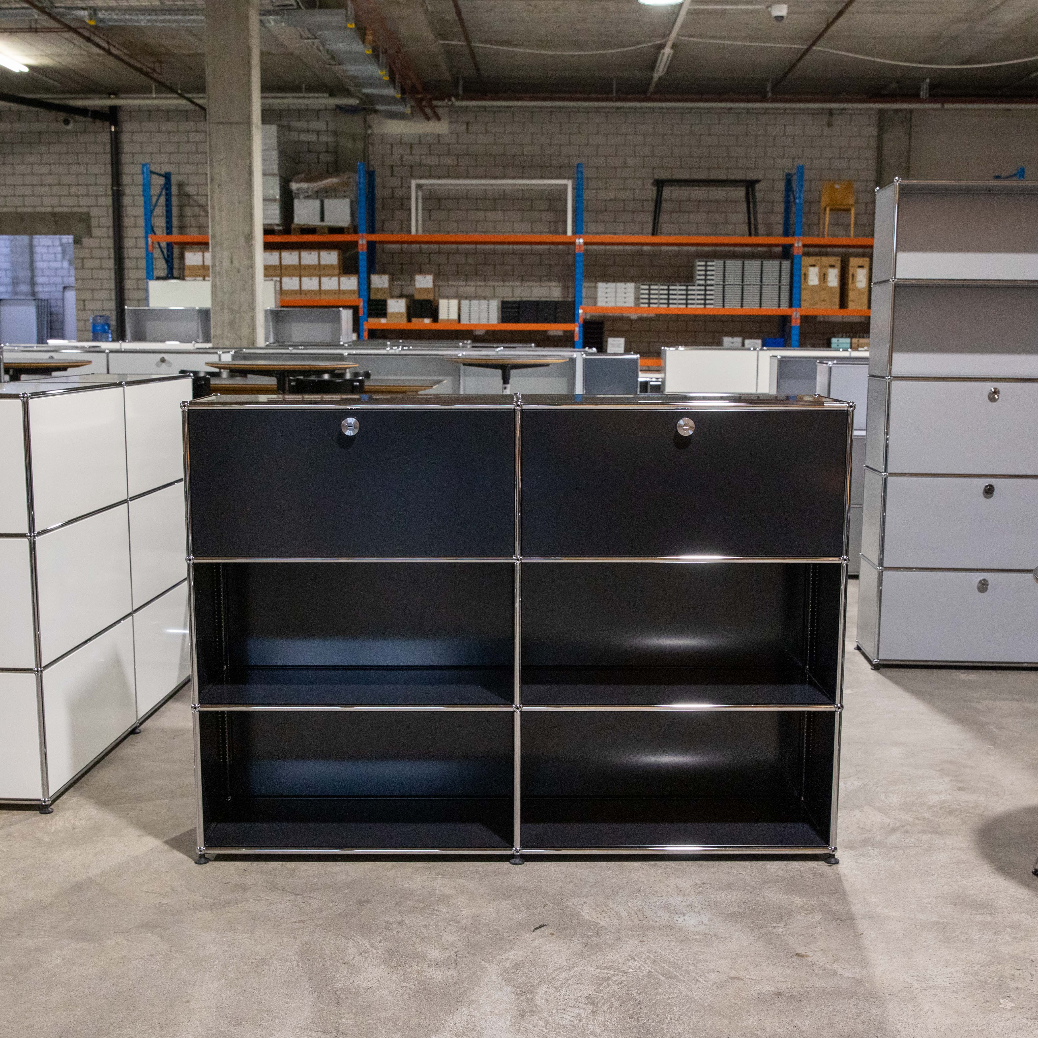 Schwarzes USM Haller Regal (2x3), mittig platziert. Oben zwei verschließbare Klapptüren, darunter zwei Ebenen mit offenen Fächern. Aus schwarzen Metallpaneelen und verchromten Rohren. Im Hintergrund weitere USM Möbel und Lagerregale.