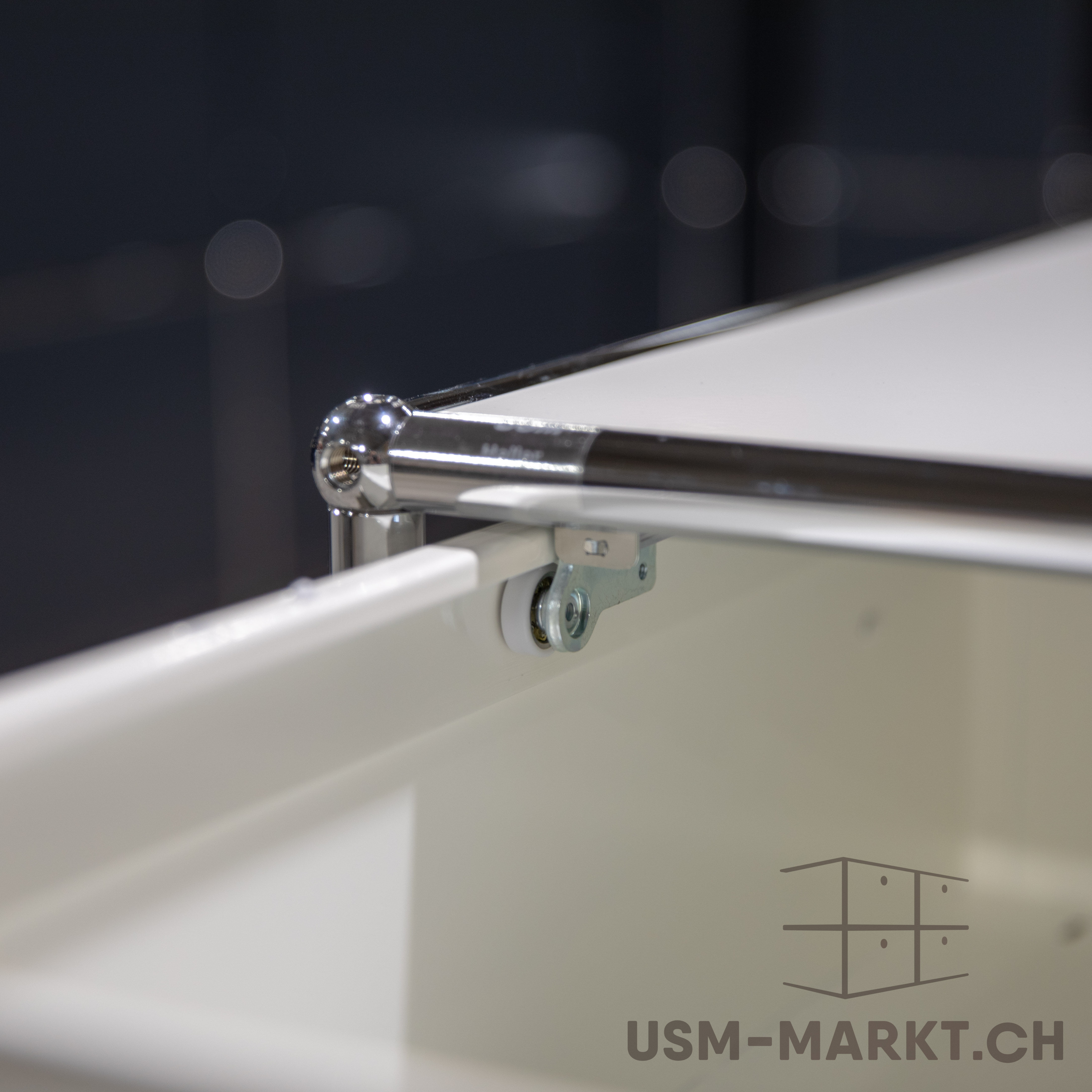 Primo piano di un elemento d'arredo bianco del sistema USM Haller, sostenuto da un'asta cromata lucida e da un giunto sferico. È visibile un piccolo rullo bianco con supporto metallico, che fa pensare a un cassetto o a un cassettone estraibile.