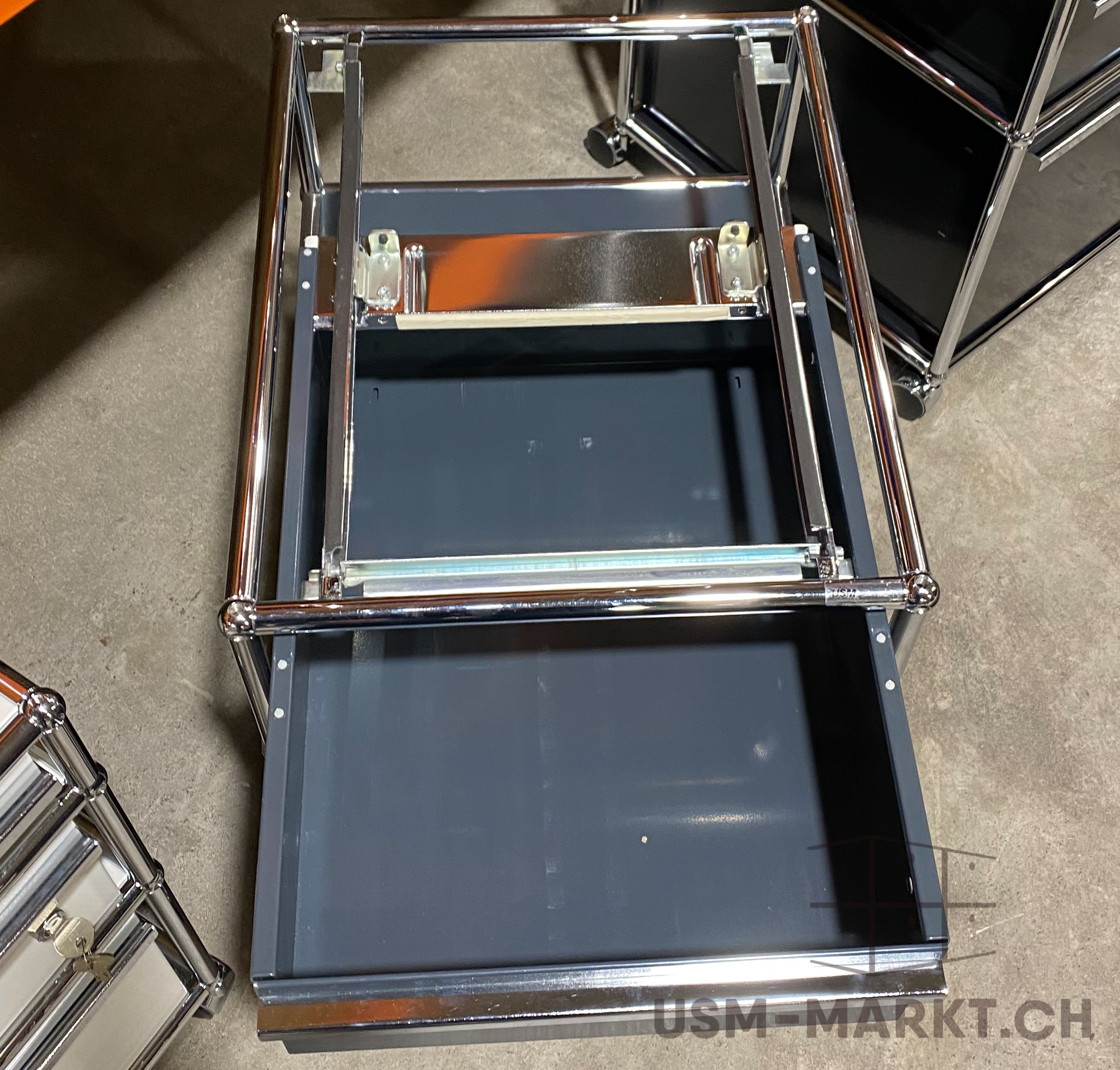 A dark grey USM Haller filing cabinet with the drawer fully extended, standing on a concrete floor. Polished chrome tubular frame; a metal hanging file frame in the open compartment. The top and side of a black cabinet in the background.