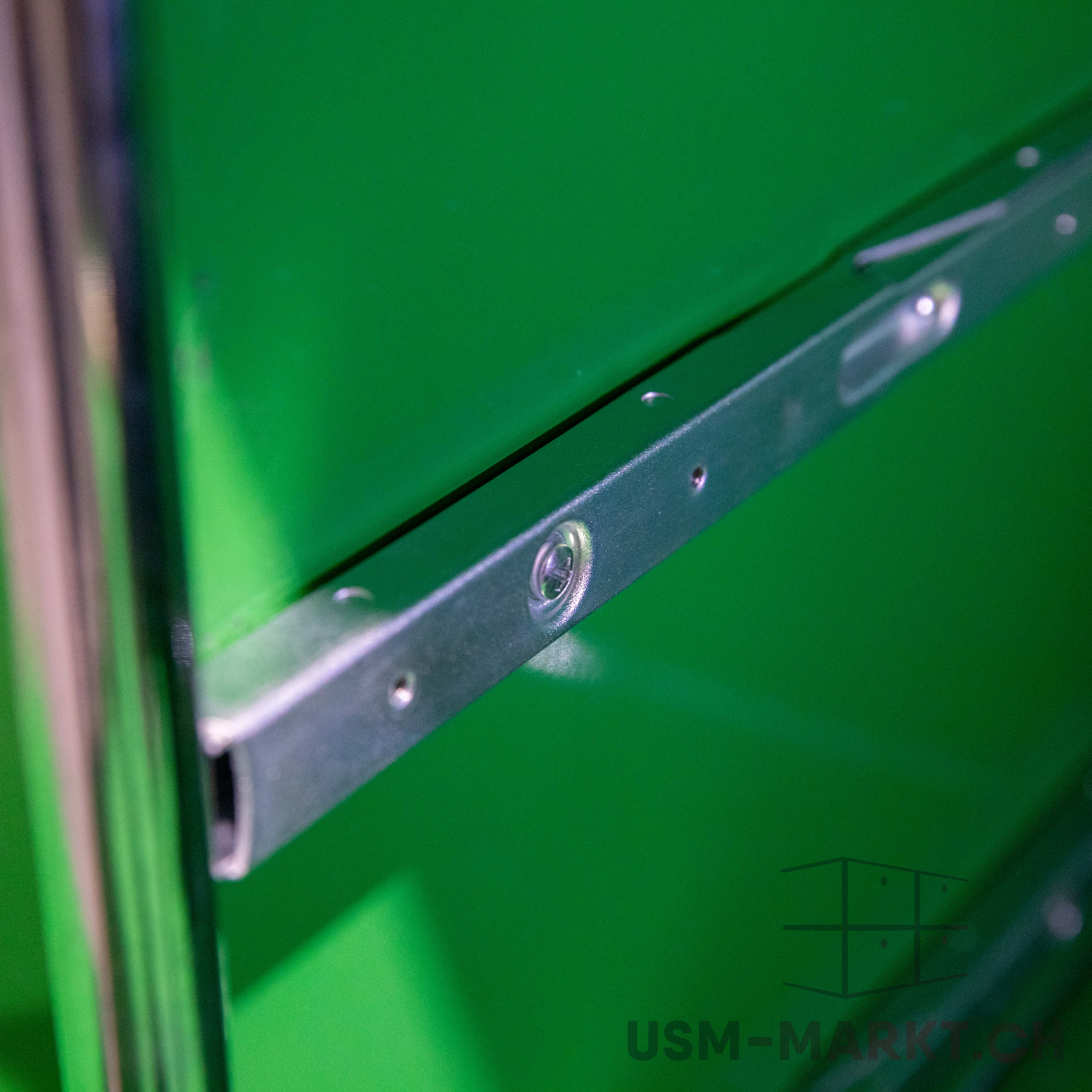 Close-up of a silver metal element running horizontally across a bright green surface. Several screw heads are visible. On the left, a blurred vertical rod. Fine textures, technical aesthetic.