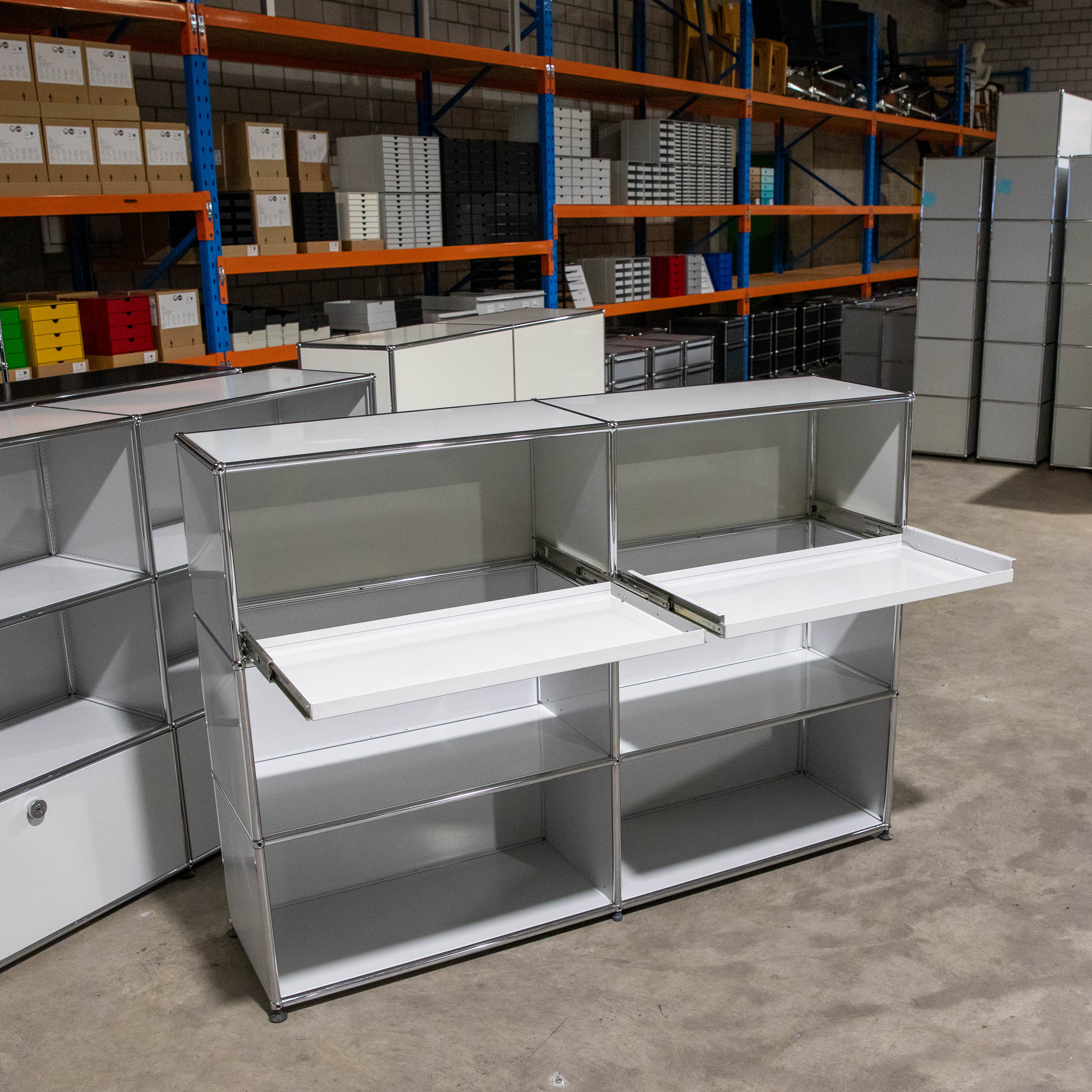 A light grey, second-hand USM Haller sideboard with a modular design, currently in stock. Three shelf heights, two widths; two central pull-out shelves are open. Further shelves and stock items in the background; condition ‘as seen’.