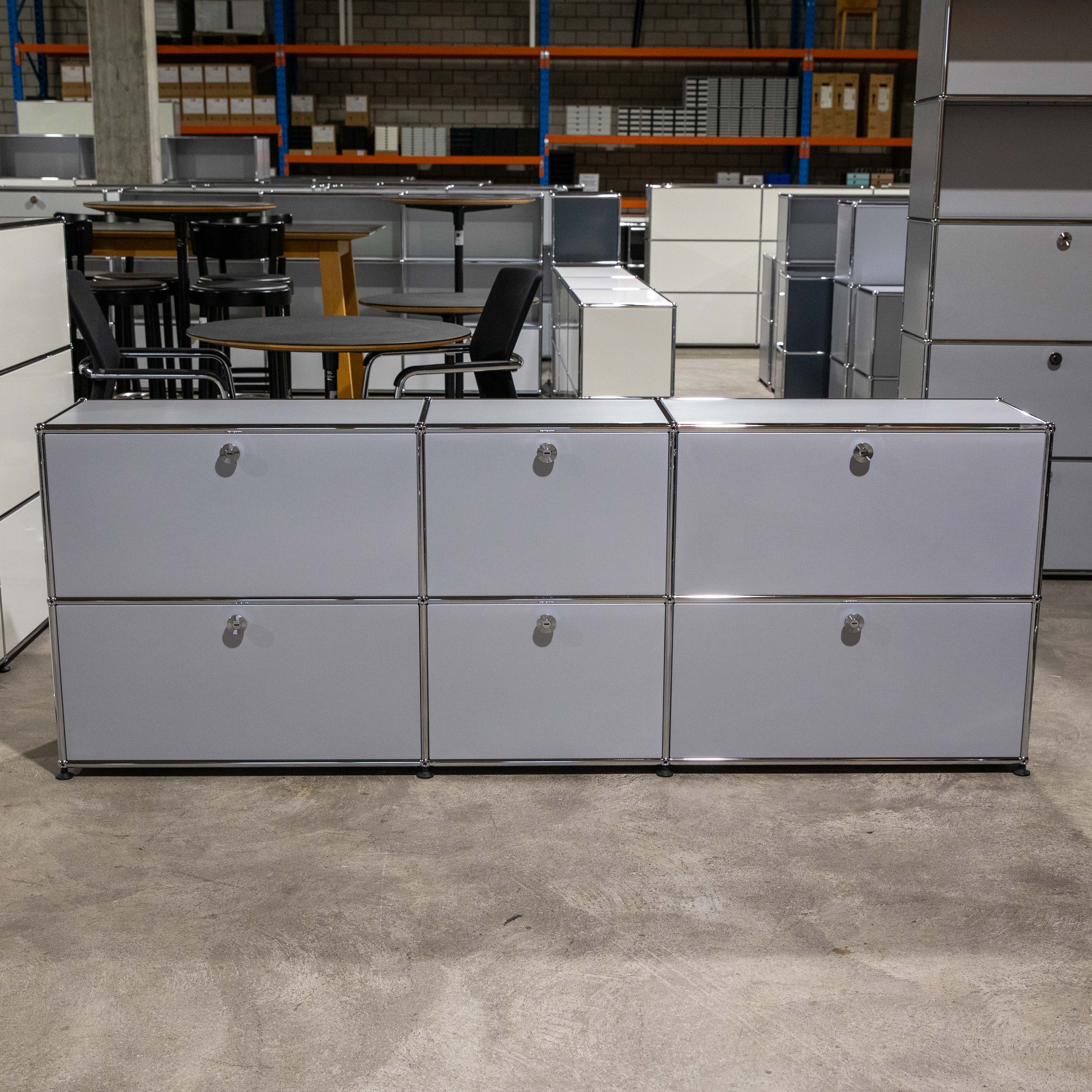 Modernes USM Haller Sideboard in Mattsilber mit sechs Fächern (2 Reihen, 3 Spalten) und Chromgriffen. Glänzende Chromstangen rahmen die Platten ein. Steht auf hellgrauem Betonboden in einer Lagerhalle.