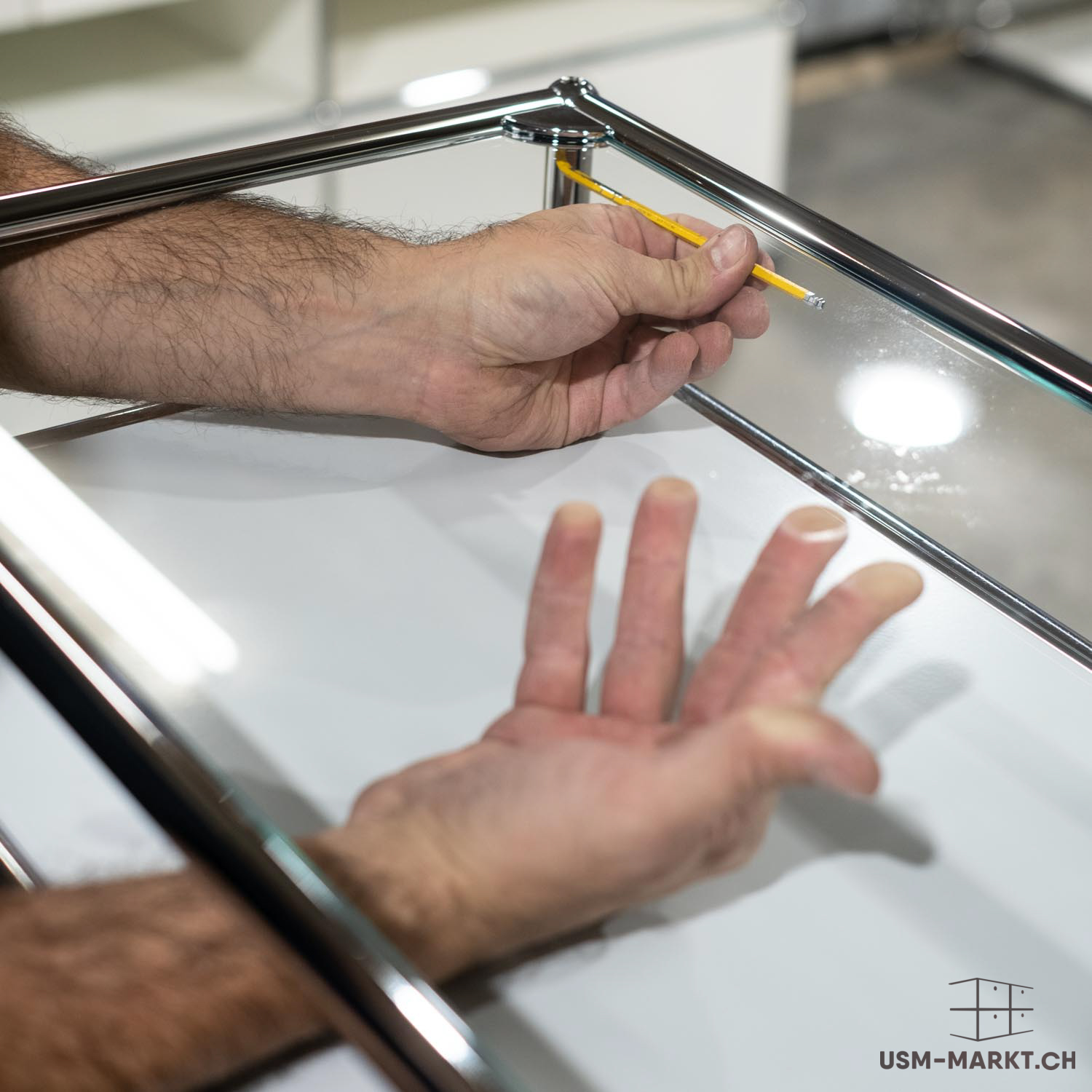 Le mani, con uno strumento giallo, posizionano un elemento trasparente in un mobile modulare di tubi cromati lucidi e superfici bianche. Il lavoro si svolge sul bordo superiore, mentre l'altra mano poggia su una superficie.