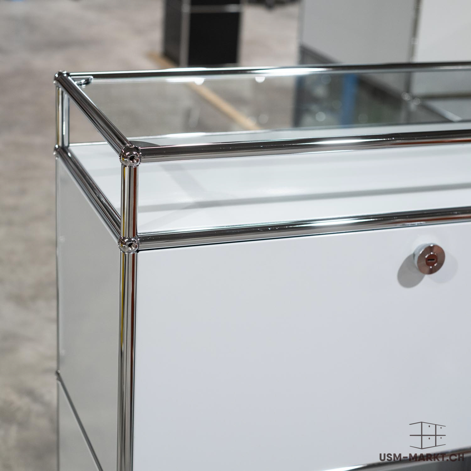 Mobile modulare bianco con tubi cromati lucidi e piano in vetro trasparente. Linee pulite e stile moderno, con un piccolo foro per la chiave sulla parte anteriore. Sullo sfondo si intravede un magazzino.