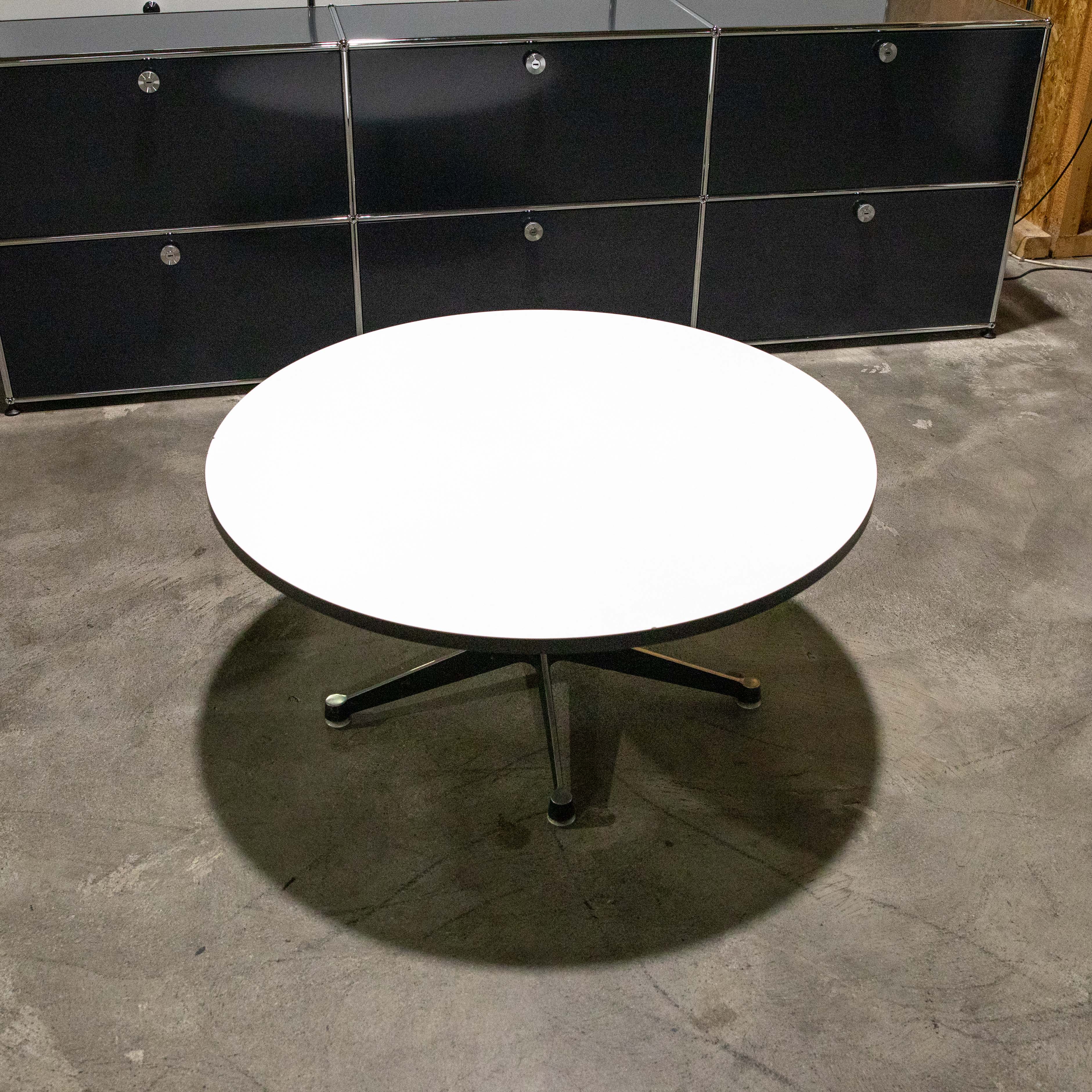 Runder, weißer Sofatisch von Charles & Ray Eames auf zylindrischem Chromfuß mit vier dunklen Beinen, mittig auf grauem Betonboden. Er wirft einen großen, runden Schatten. Dahinter schwarzes Modulregal mit sechs Fächern und silbernen Rahmen.