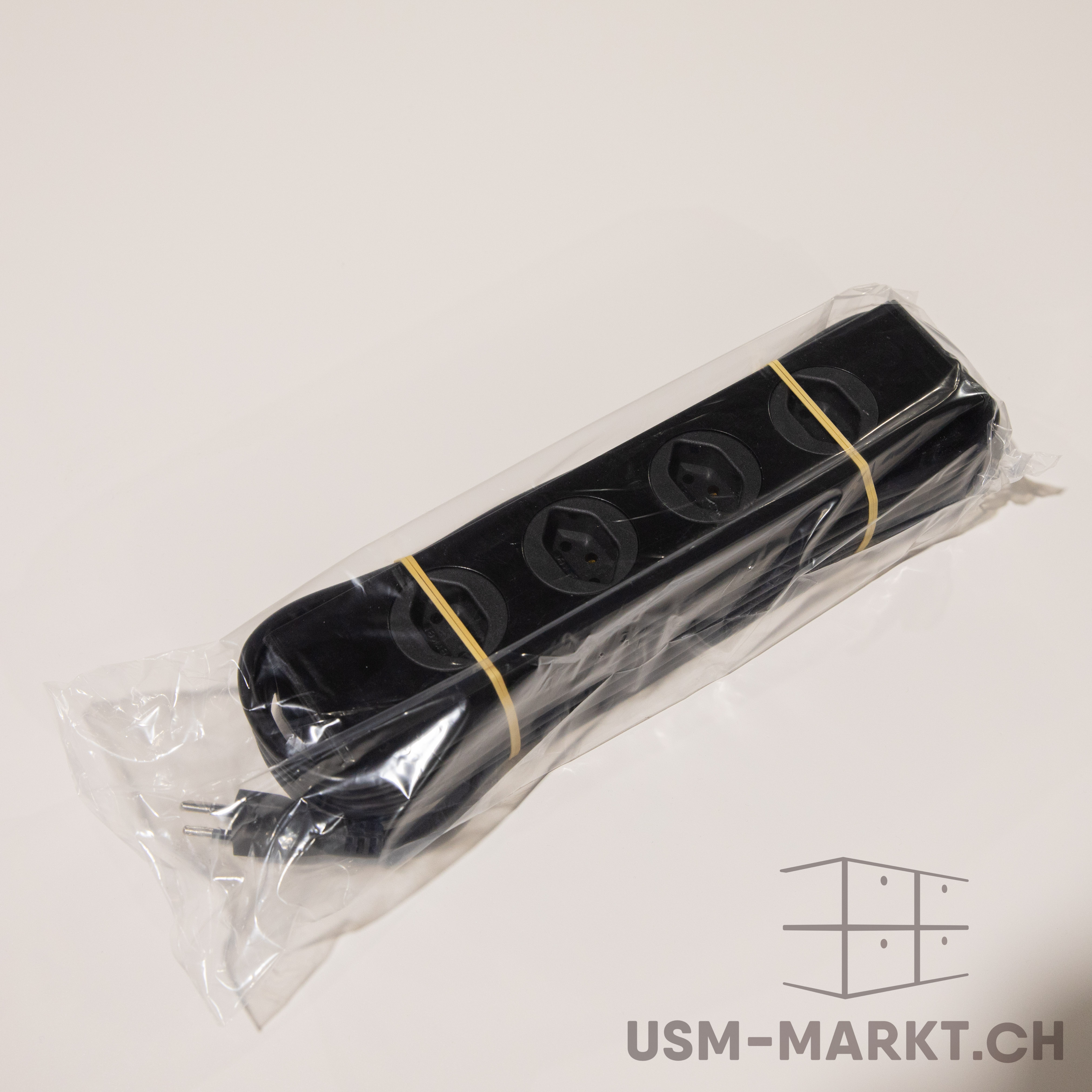 A black power strip with four Schuko sockets, wrapped in transparent plastic film and secured with two yellow rubber bands. The black power plug is visible on the left. It lies diagonally on a light-coloured background.