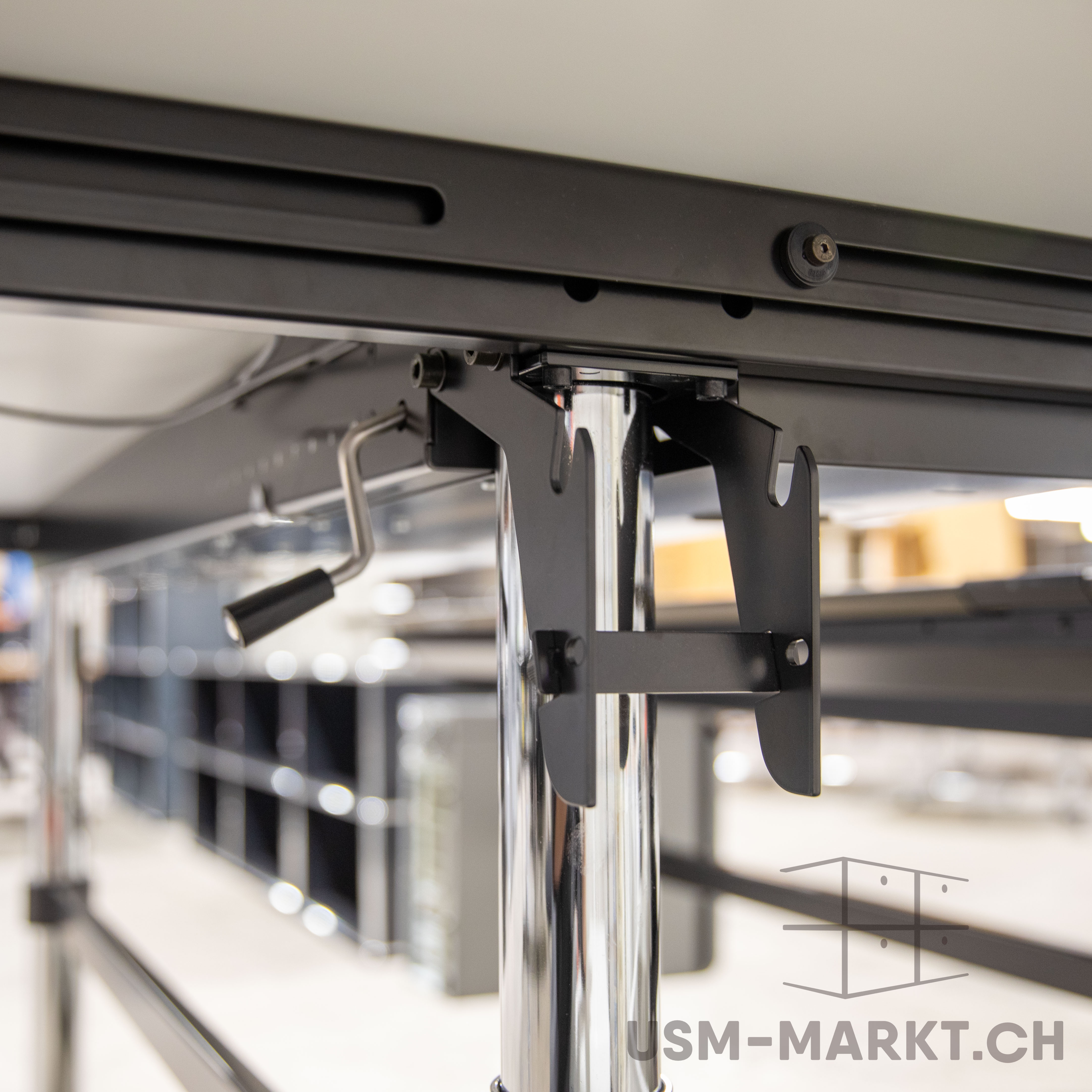 Nahaufnahme der Unterseite eines höhenverstellbaren Tisches: schwarzer Metallrahmen an heller Platte, über schwarze Halterung mit glänzend verchromtem Tischbein verbunden. Links ein Kurbelgriff zur Höhenverstellung, Hintergrund unscharf.