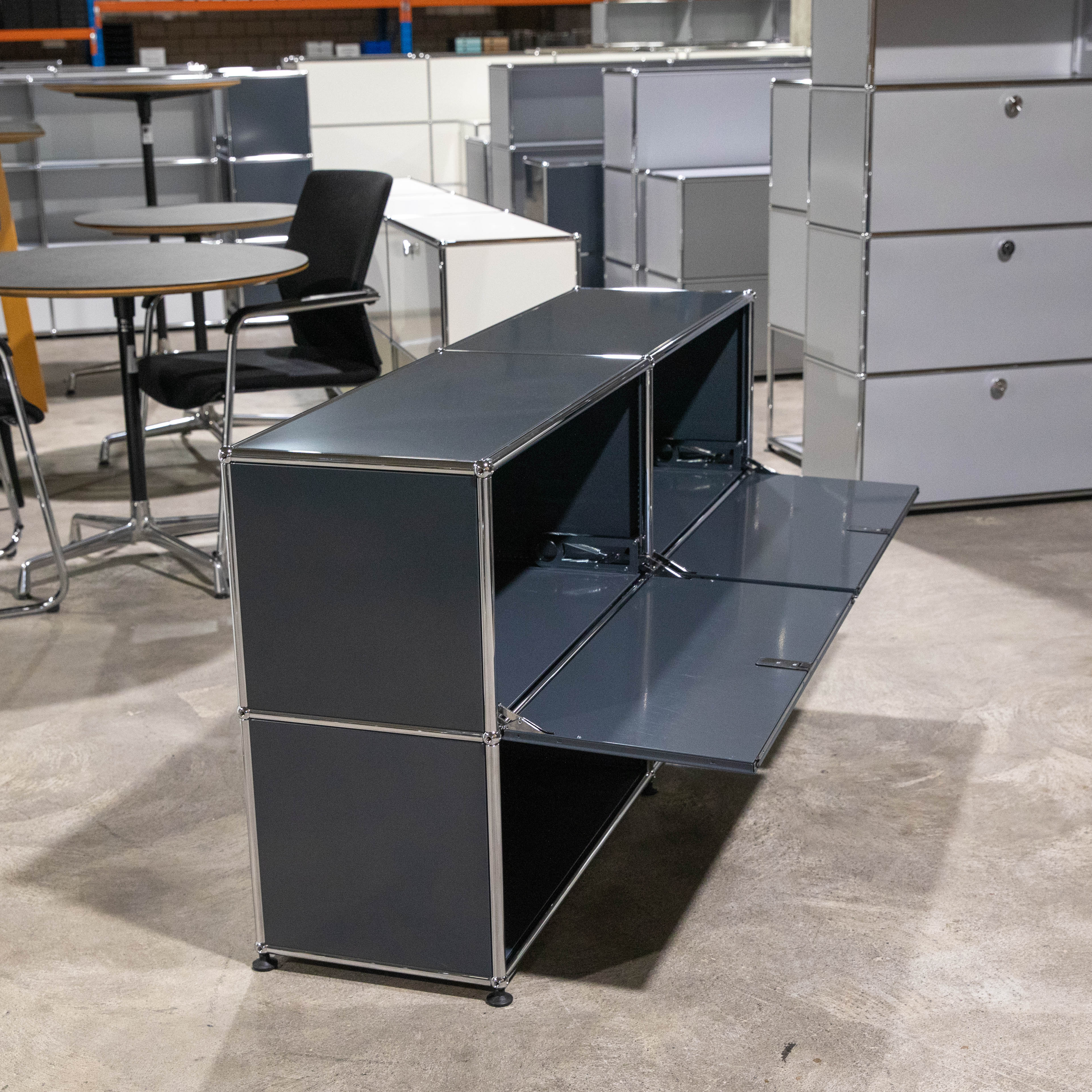USM Haller Sideboard in Anthrazit, zwei Felder breit, zwei Fächer hoch. Obere und untere rechte Klapptür sind geöffnet und zeigen die Innenfächer des gebrauchten Möbels mit glänzenden Chromstreben. Im Hintergrund Büroregale und Tische im Lagerraum.