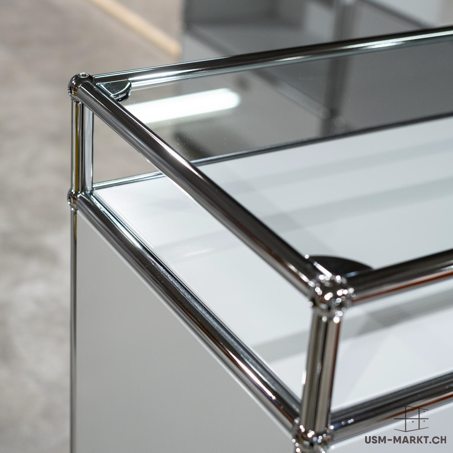 Primo piano di un mobile modulare moderno con tubi metallici cromati lucidi che ne costituiscono la struttura, collegati da giunti sferici. Pannelli bianchi opachi e un ripiano superiore in vetro trasparente che riflette la luce.