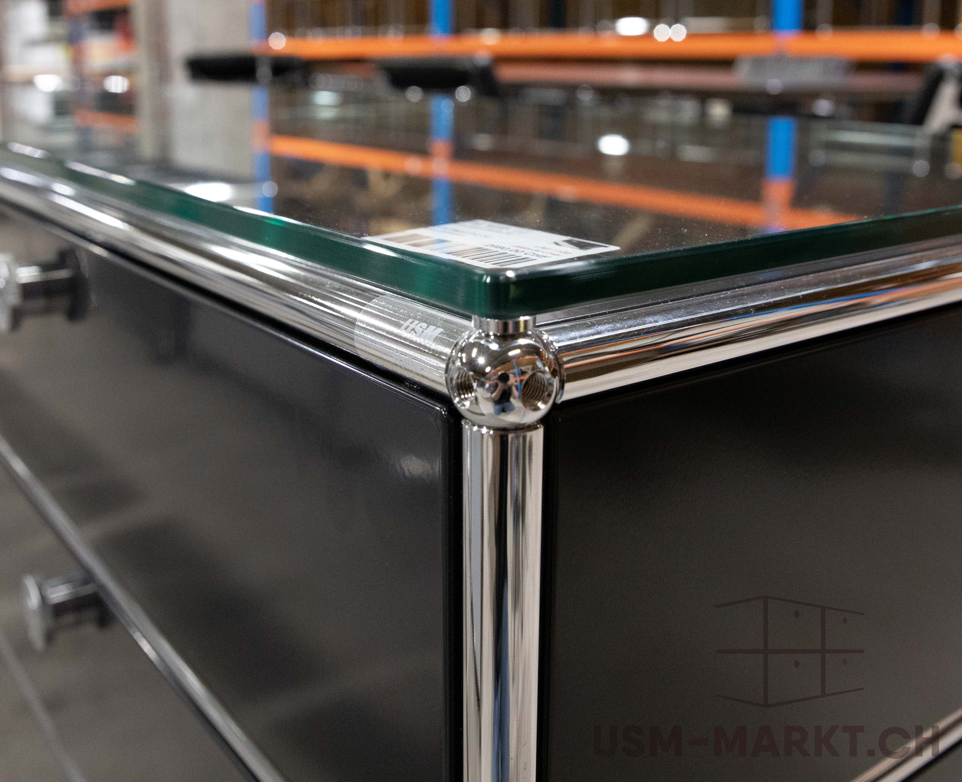 A close-up of a modular piece of furniture in dark grey with polished chrome tubes and ball connectors, topped with a clear glass top. The surface is slightly reflective, whilst orange and blue storage shelves are visible in the blurred background.