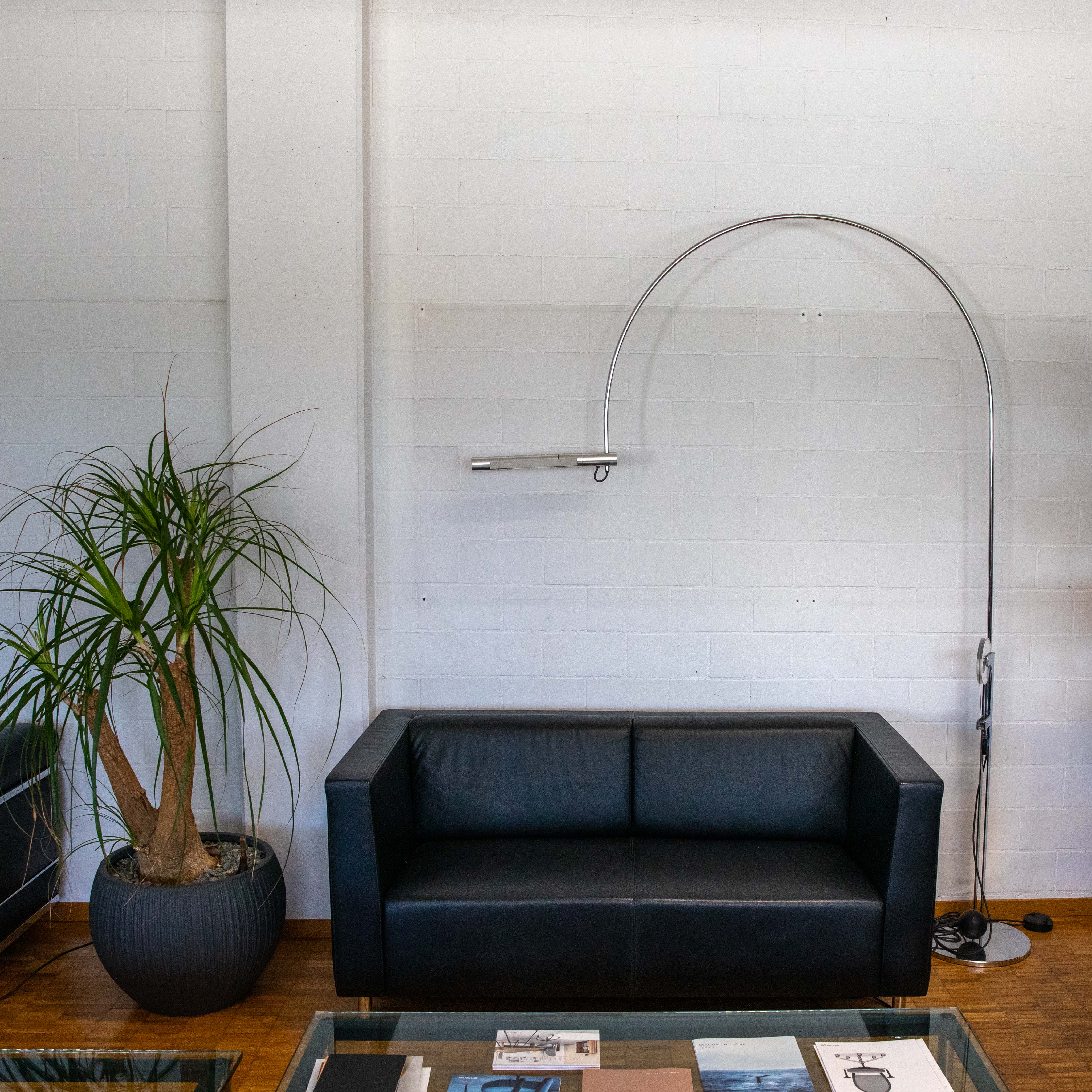 Lampada ad arco moderna "Baltensweiler" argento, con braccio metallico sottile e ricurvo e testa alogena orizzontale. Vicino a un divano in pelle nera e a una grande pianta in vaso verde, davanti a parete di mattoni bianchi, su caldo pavimento in legno.