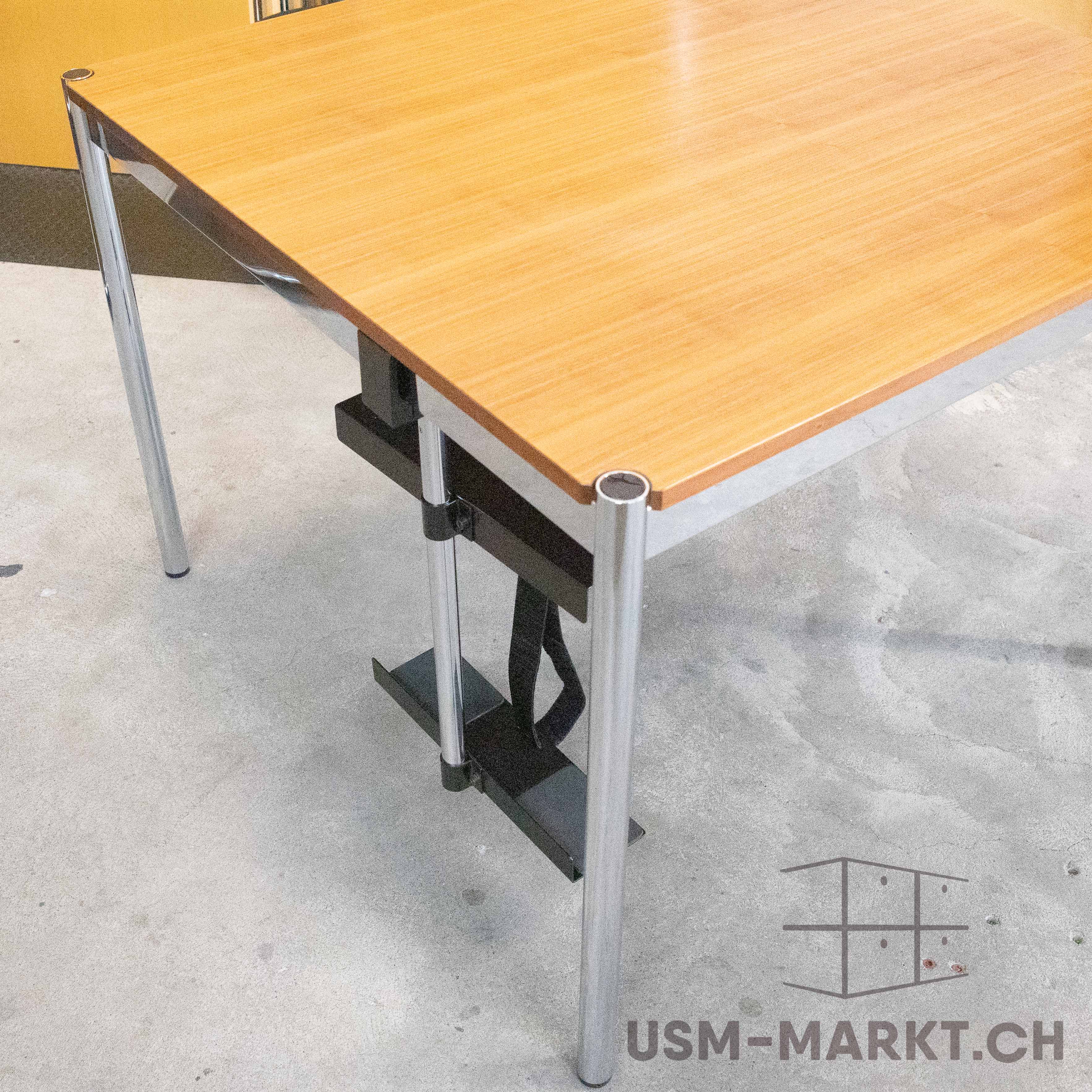 A modern desk with a light-coloured wooden top featuring a distinct grain, supported by shiny chrome legs. A black metal bracket is fitted beneath the top. Set against a grey concrete floor, with a yellowish wall in the background.