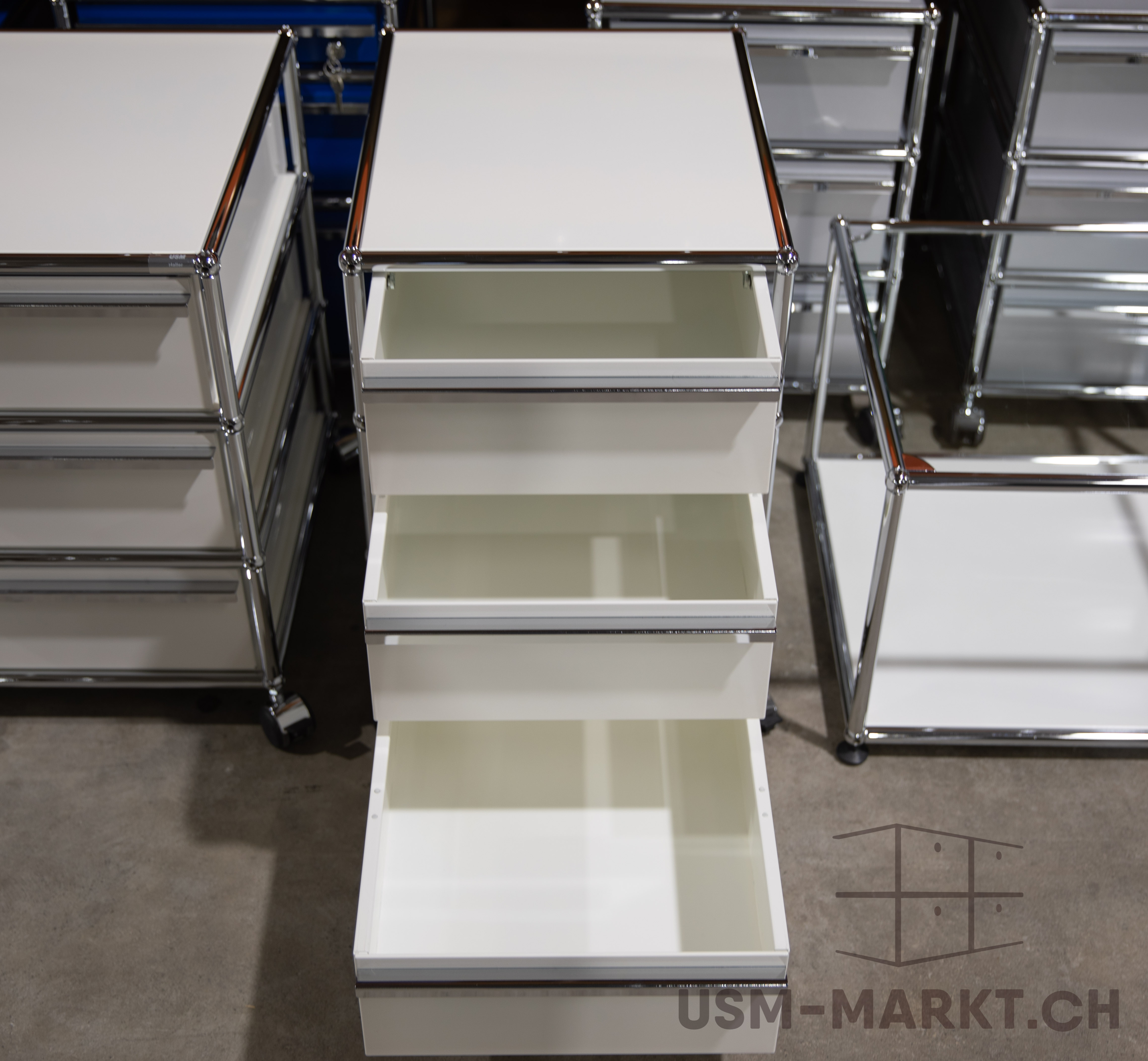 Contenitore su ruote USM Haller alto, bianco, con 4 cassetti: superiore leggermente aperto, secondo e terzo semiaperti, inferiore completamente aperto con interno vuoto visibile. Pannelli bianchi e telaio cromato, su pavimento in cemento.