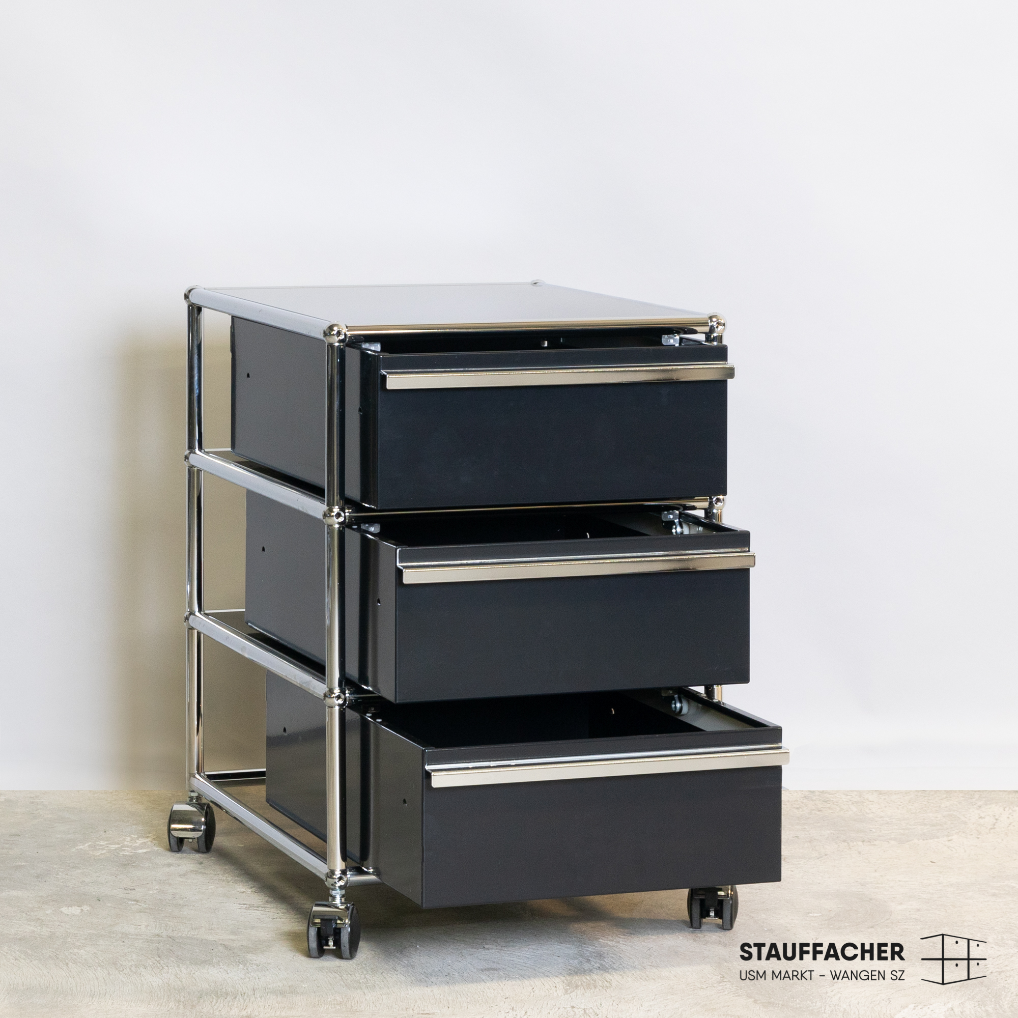 Ein schwarzer, modularer Rollcontainer mit drei übereinanderliegenden Schubladen aus Metall, die jeweils leicht geöffnet sind und silberne Griffe besitzen. Er steht auf einem hellen Betonboden vor einer weißen Wand und hat kleine, dunkle Rollen.