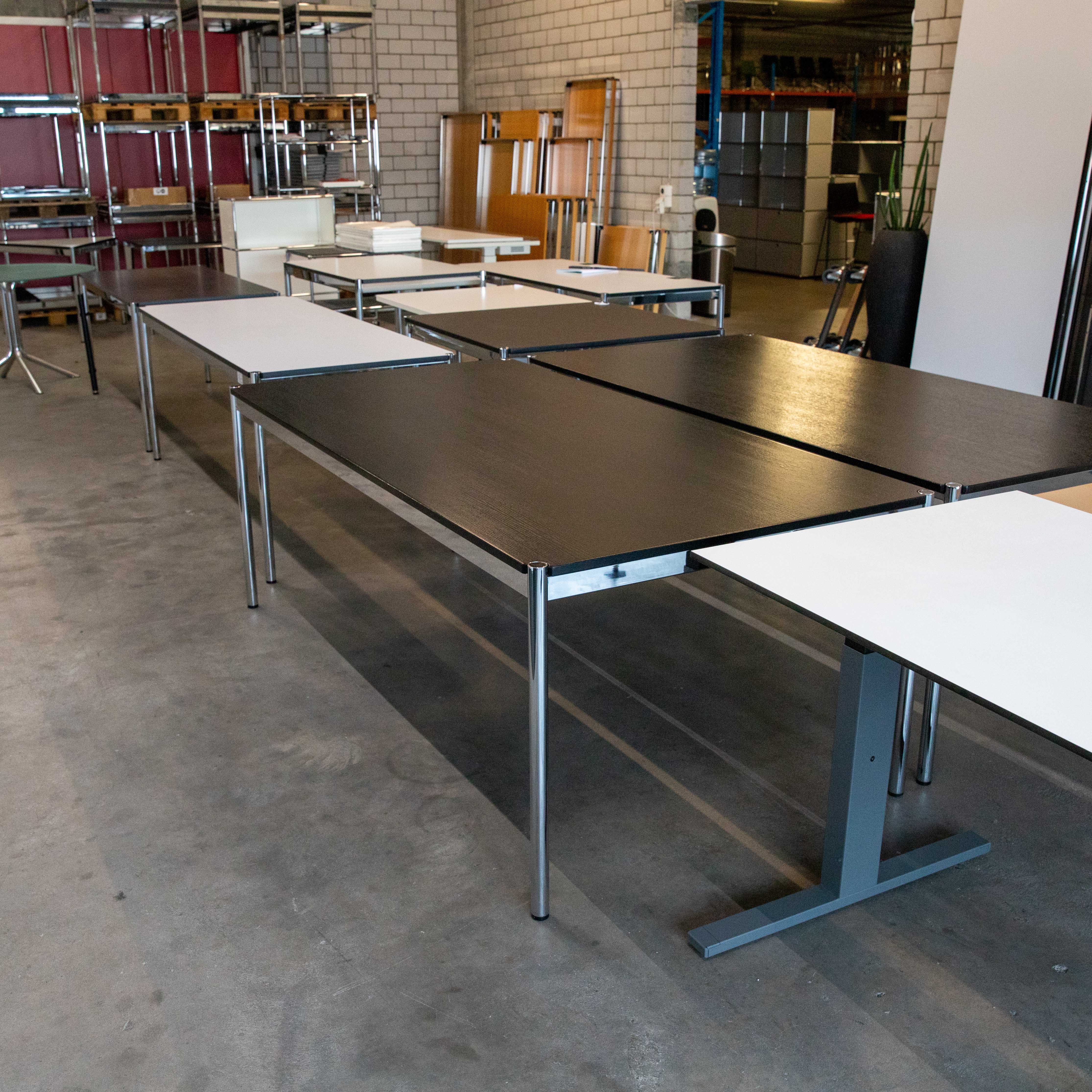 Plusieurs tables USM Haller d'occasion, aux pieds chromés brillants et plateaux variés, sont sur un sol en béton. À l'avant, plateaux en chêne noir et blancs. À l'arrière, rangements et autres meubles.