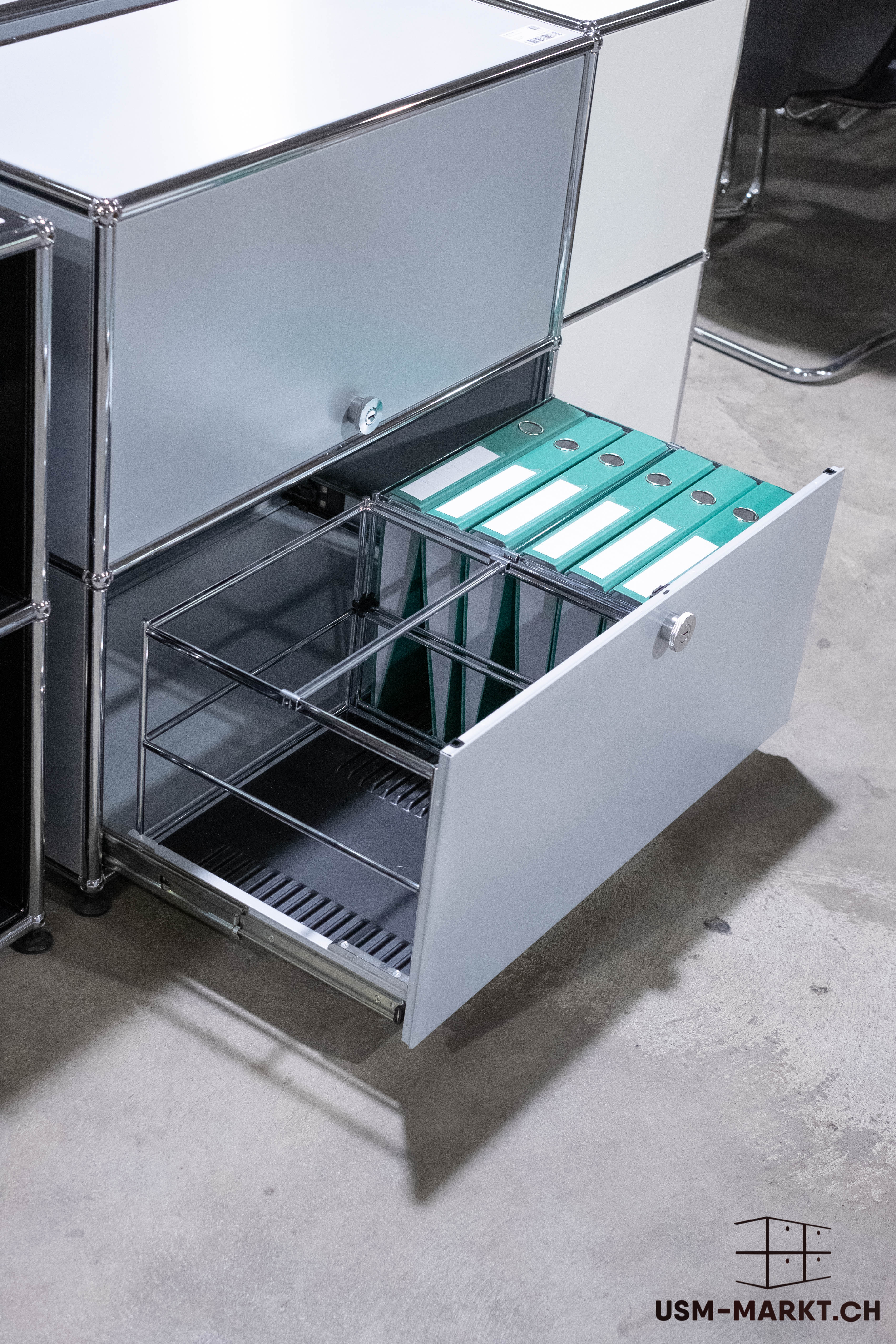A silver USM Haller filing cabinet with a wide-open bottom drawer. The drawer contains several mint-green file folders, which stand vertically within a metal frame. The cabinet stands on a grey concrete floor.