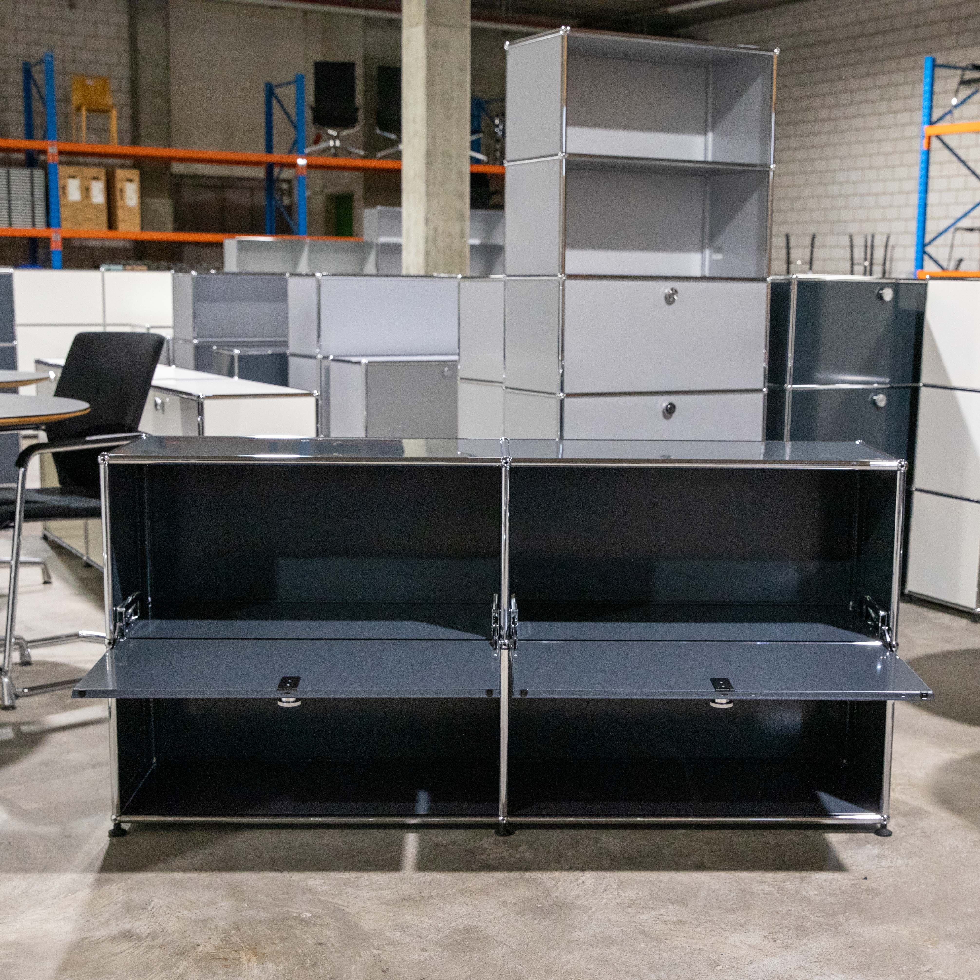 Anthrazitfarbenes USM Haller Sideboard, 2 Felder breit und 2 Fächer hoch, mittig im Raum positioniert. Beide unteren Klapptüren sind geöffnet und zeigen das dunkle Innere. Im Hintergrund modulare Möbel und Industrieregale.