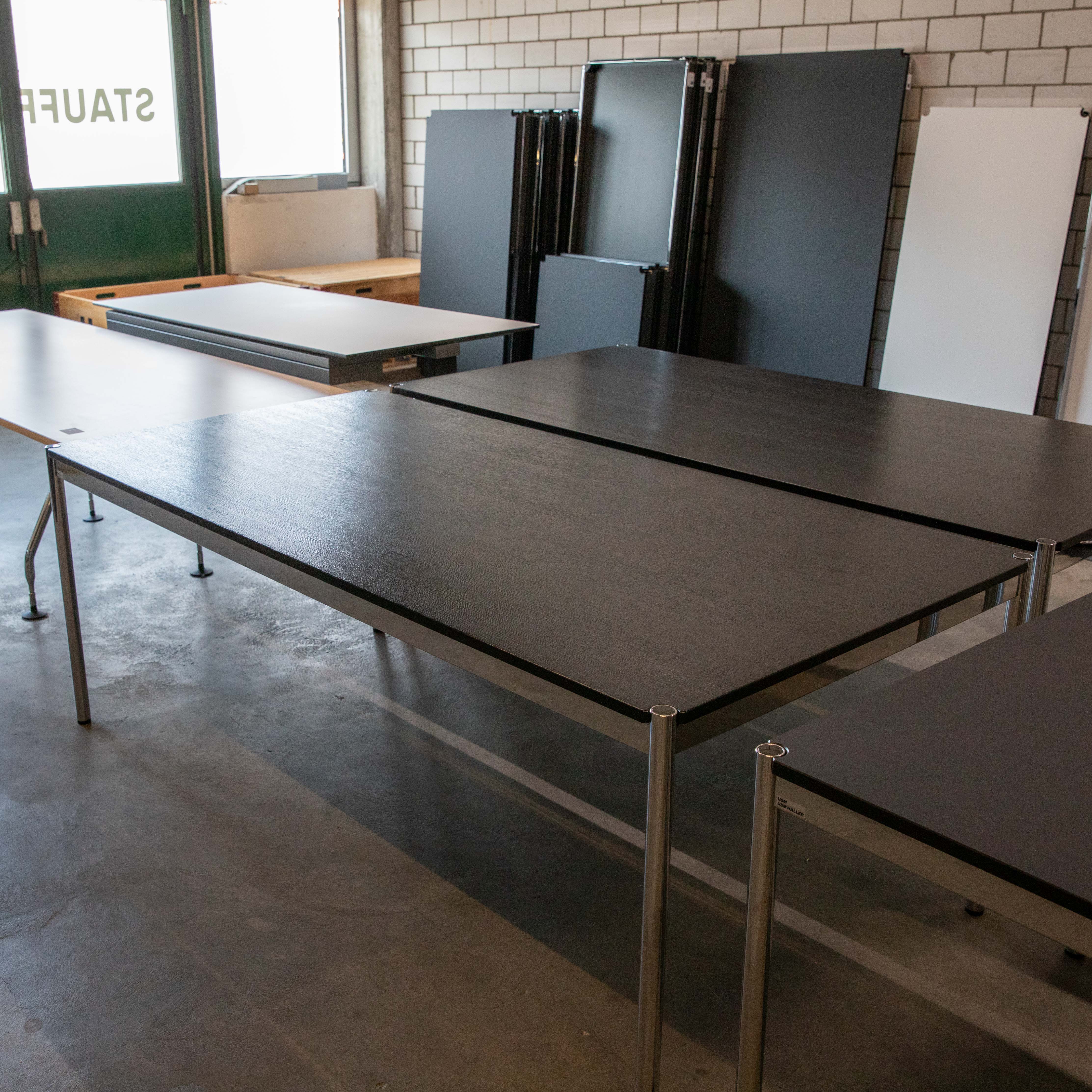 A large, rectangular USM Haller table with a dark brown-black wood-grain tabletop and polished chrome legs on a concrete floor. In the background, there are further tables and stacked black tabletops against a brick wall.