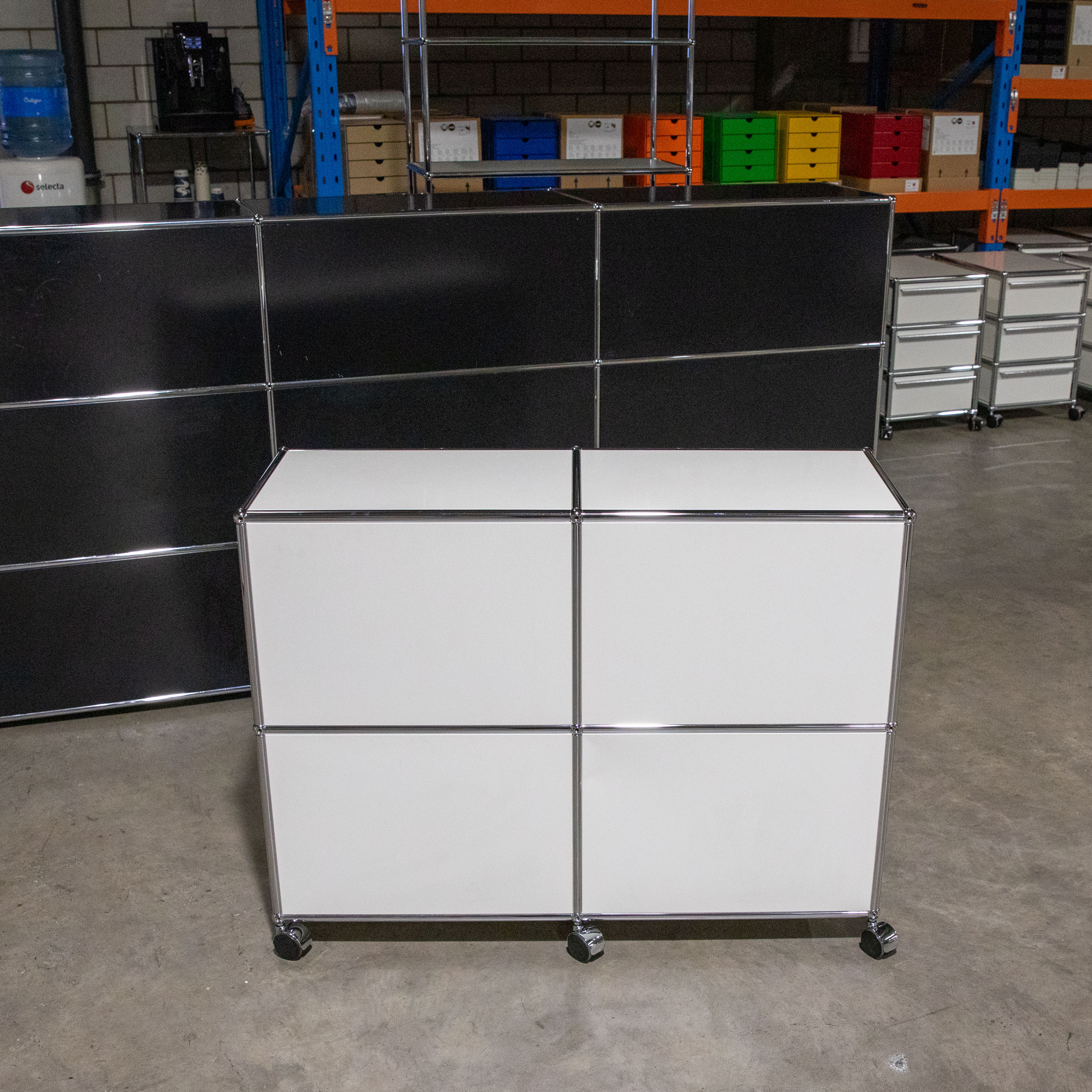 Weißes USM Haller Möbel aus vier Modulen in 2x2 Anordnung auf Rollen. Modulare Konstruktion mit mattweißen Elementen und glänzenden, verchromten Stahlrohren und -verbindern. Im Hintergrund schwarze Möbel und farbige Lagerregale.
