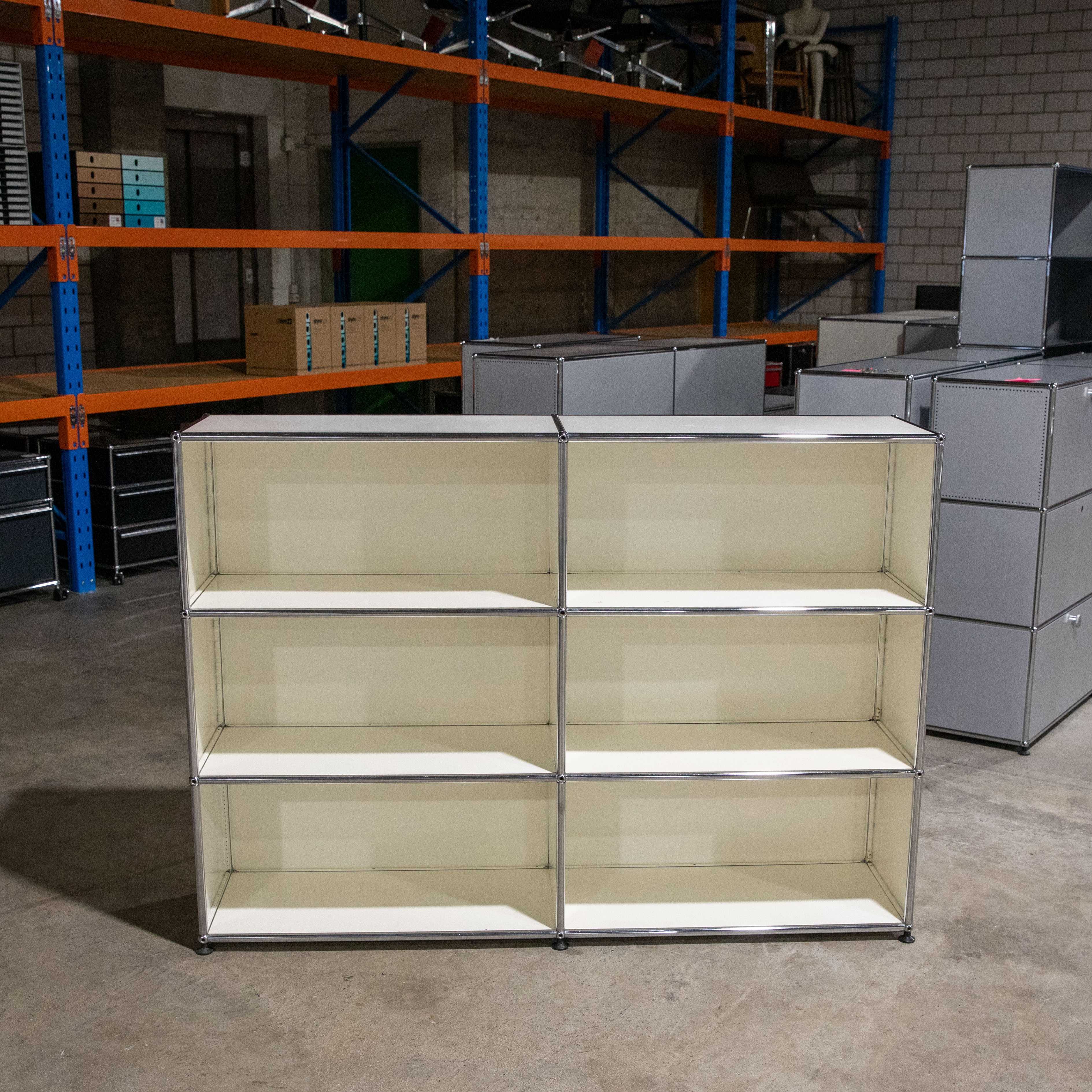 Ein beiges, dreireihiges und zweispaltiges offenes Regalsystem mit glänzenden Chromrahmen steht auf hellgrauem Betonboden in einem Lagerraum. Im Hintergrund sind orange-blaue Metallregale mit Kartons und Bürostühlen sowie Stapel grauer Schrankelemente.