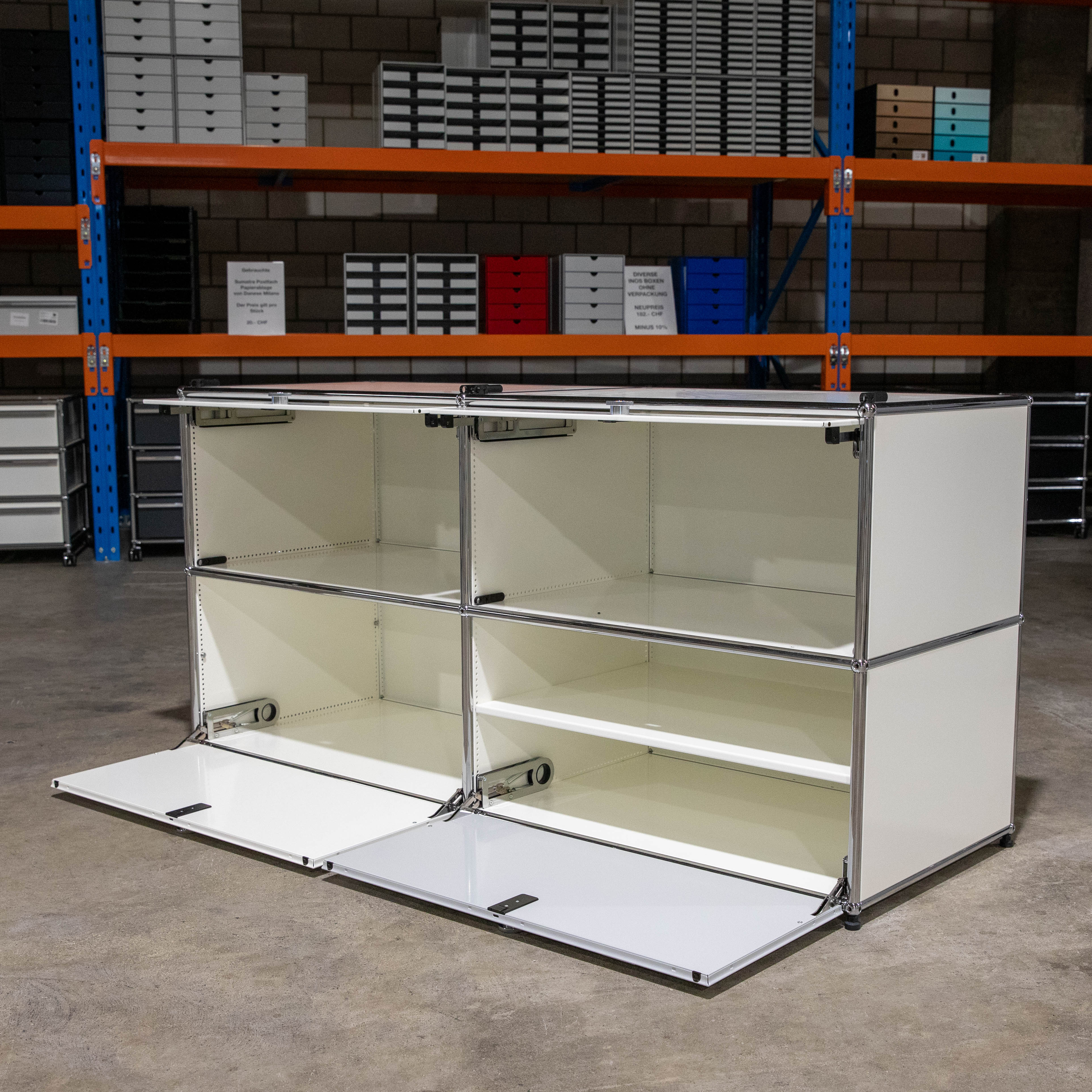 Weiß-lichtgraues USM Haller Sideboard mit vier Fächern im 2x2-Raster. Modulare Konstruktion aus verchromten Stahlrohren und hellen Paneelen. Untere Klapptüren geöffnet mit Mechanik. Oben offen, rechts mit Zwischenboden.