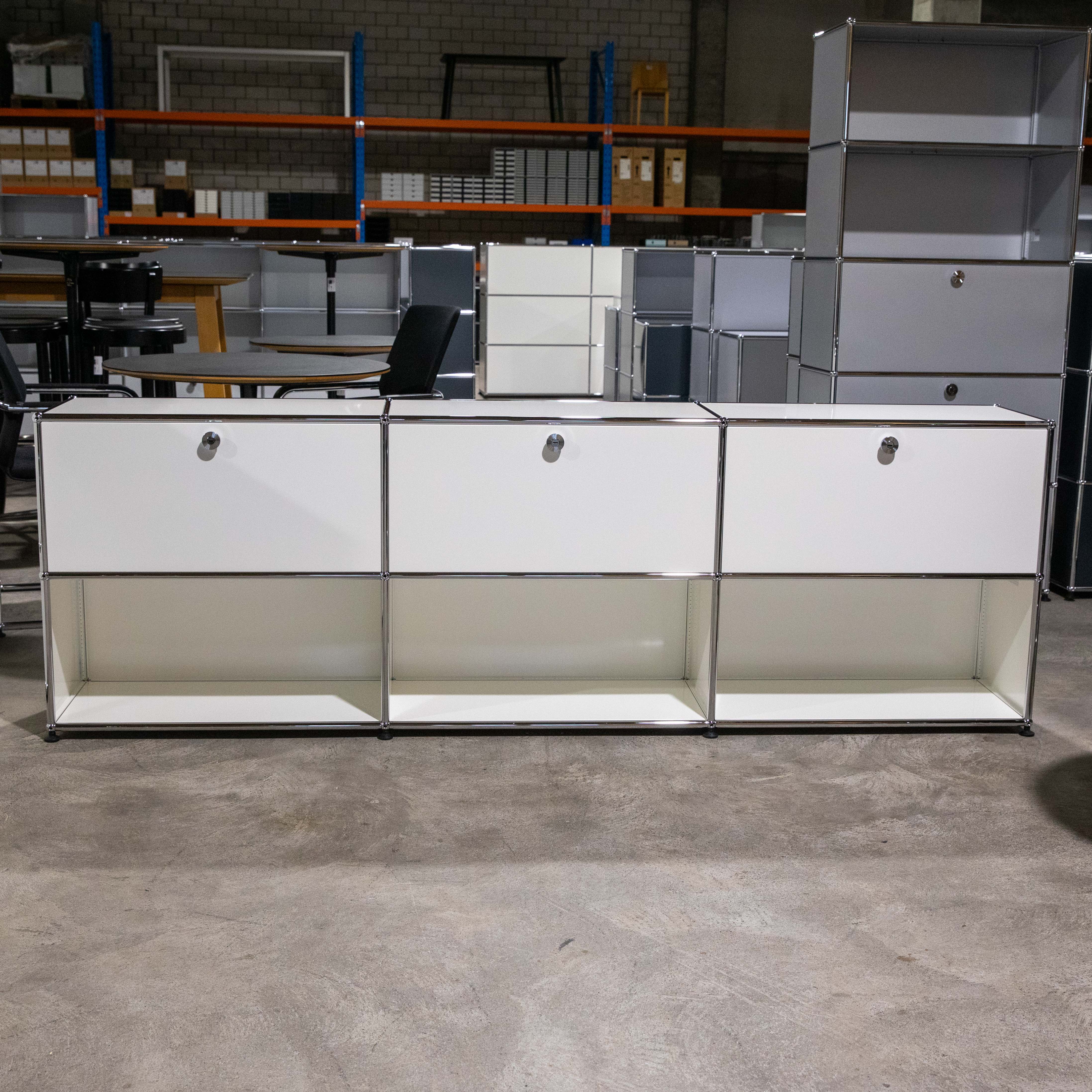 Weißes USM Haller Sideboard mit drei geschlossenen Klappfächern oben und drei offenen Fächern unten, auf grauem Betonboden. Modulare Bauweise mit verchromten Verbindungsrohren sichtbar. Im Hintergrund weitere USM Möbel und Lagerregale.