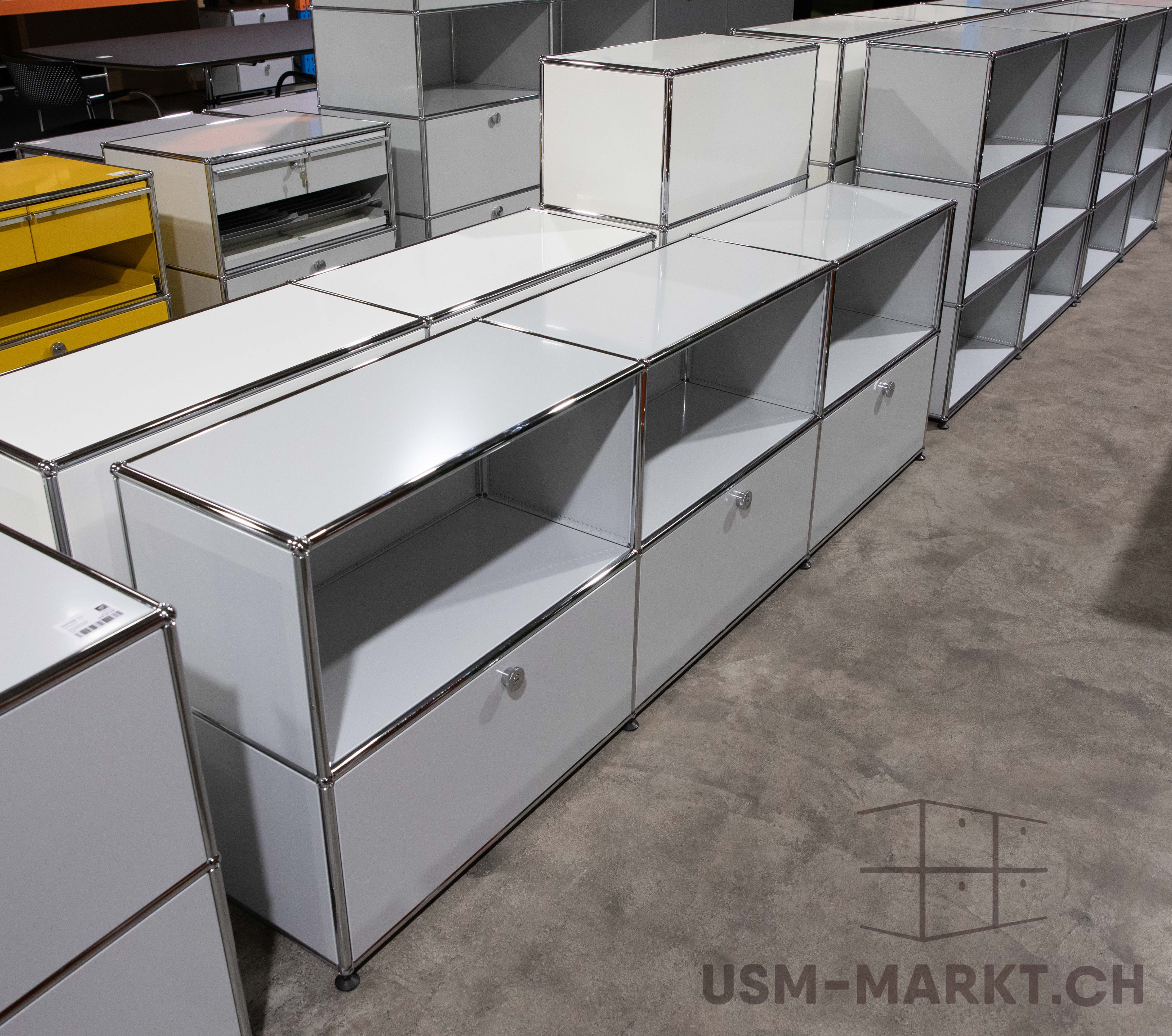 Viele modulare USM-Haller-Möbel in Lager oder Showroom. Vorn lange, hellgraue Sideboards mit glänzenden Chromrahmen, teils mit Schubladen, teils mit offenen Regalfächern. Hinten hohe Regale, gelber Rollcontainer und dunkle Bürotische.