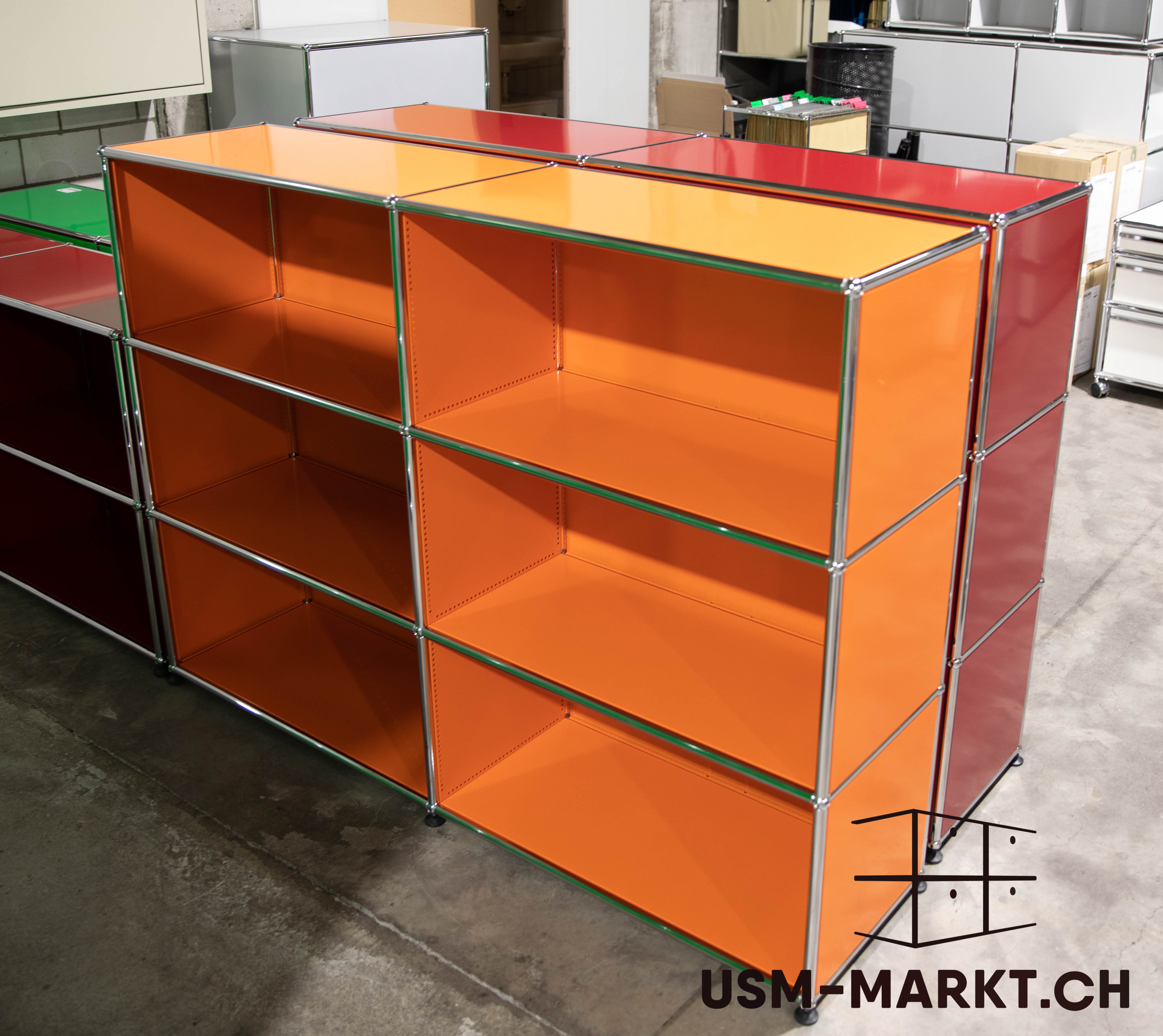 A three-tier, two-column, open shelving unit from the USM Haller system, made of bright orange metal panels and chrome rods. On the left, modules in dark red and green; on the right, in red. Set against a light grey concrete floor in the storeroom.