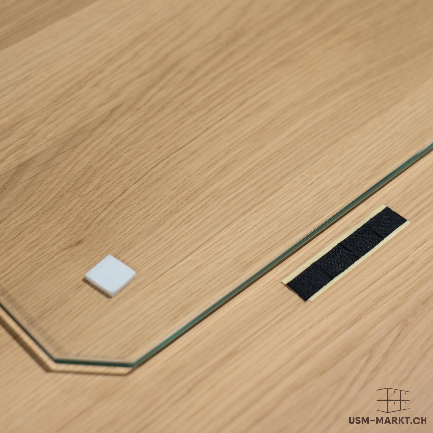 Piastra in vetro trasparente con angolo smussato su una superficie in legno marrone chiaro con venature a vista. Sul vetro è posizionato un piccolo cuscinetto quadrato bianco che funge da distanziatore. Accanto ad esso, una striscia adesiva nera.