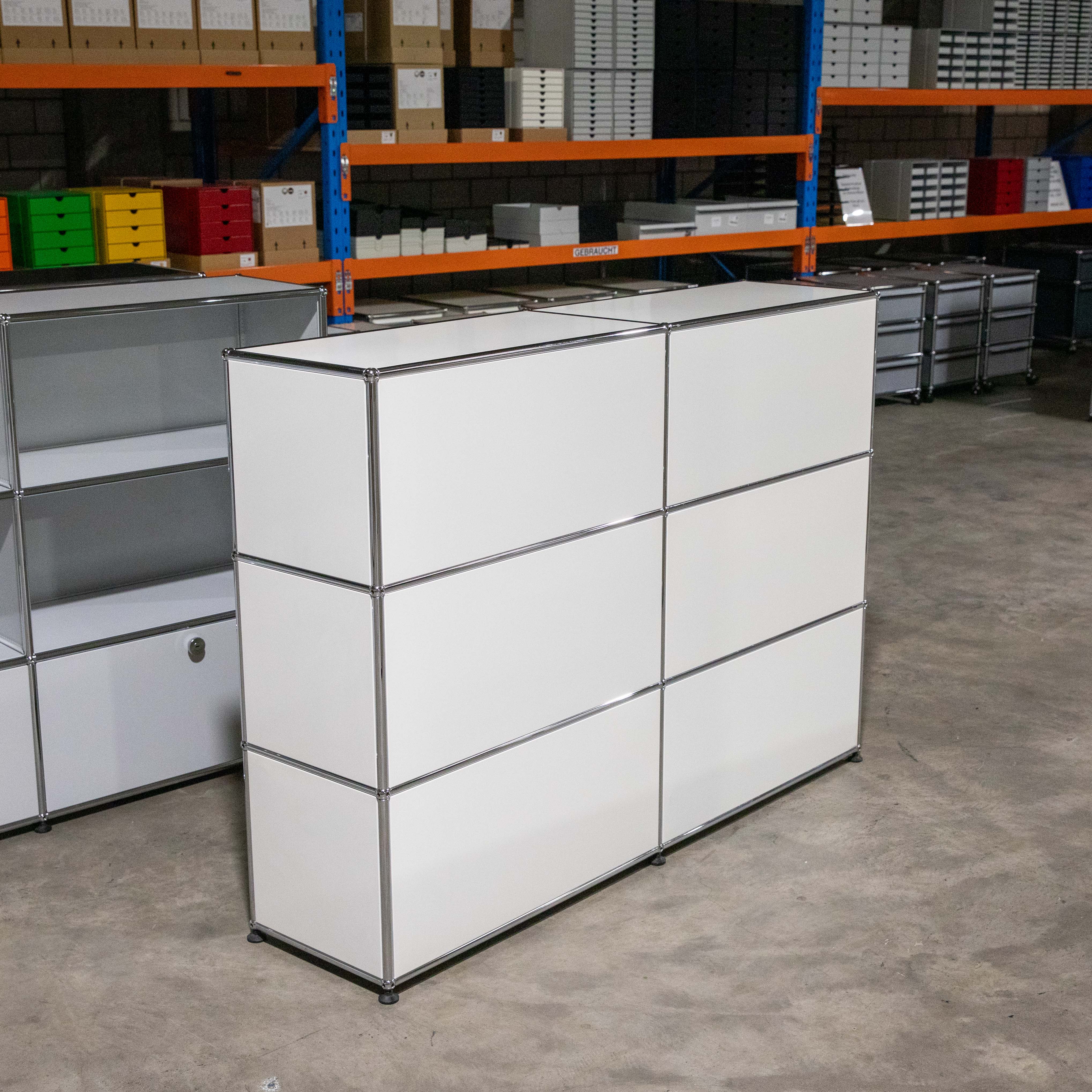 Weißes USM Haller Regal mit sechs geschlossenen Fächern (3 Reihen, 2 Spalten) auf Betonboden in einem Lager. Modulare Konstruktion mit glänzenden Chromrahmen. Hinten orange-blaue Regale mit Boxen, links teils offenes Regal.