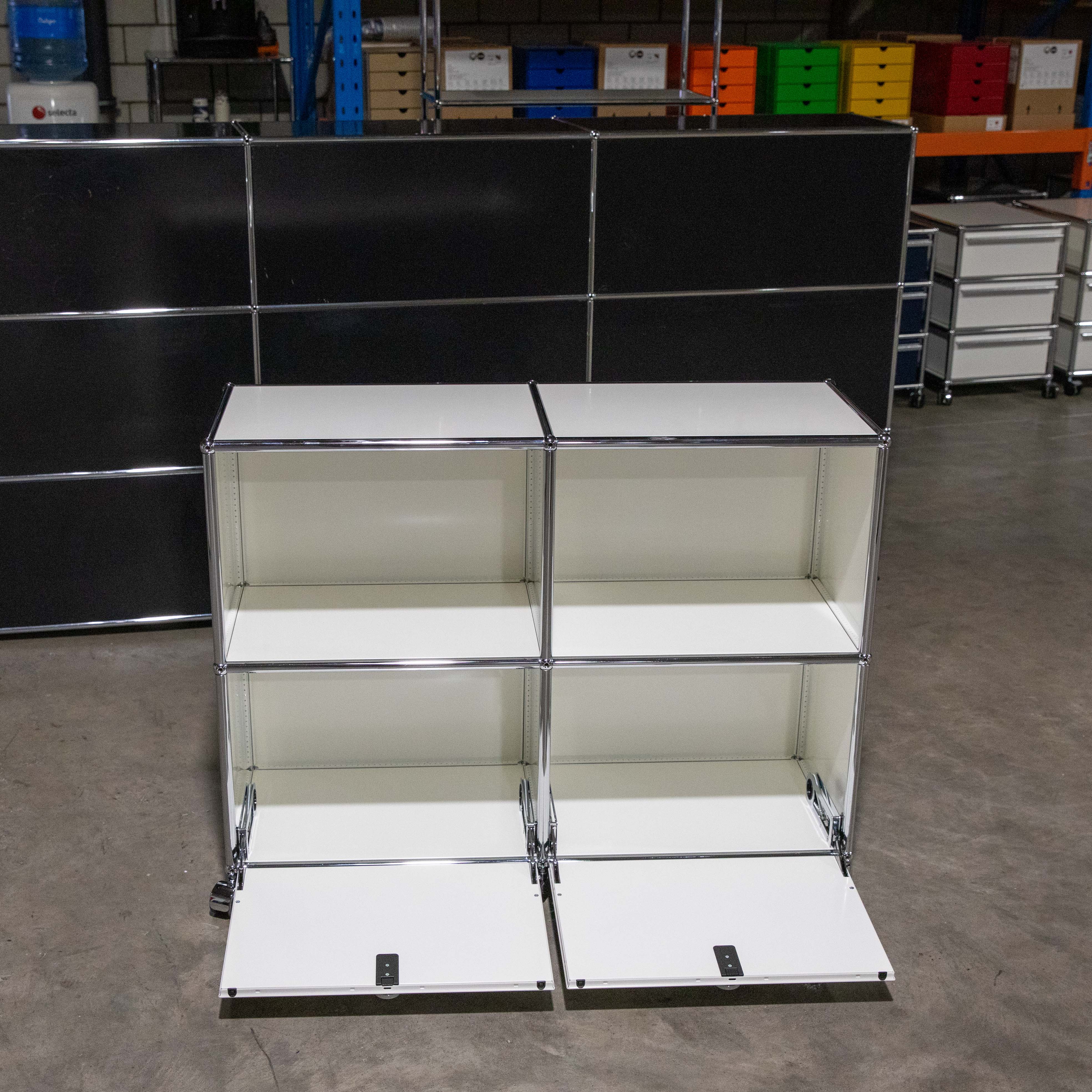 Weisses USM Haller Regal 2x2 Module auf Rollen, beide unteren Klapptüren geöffnet. Silberne Chromstreben und -verbindungen gut sichtbar. Im Hintergrund ein Lagerraum mit größerem schwarzem USM Möbelstück.