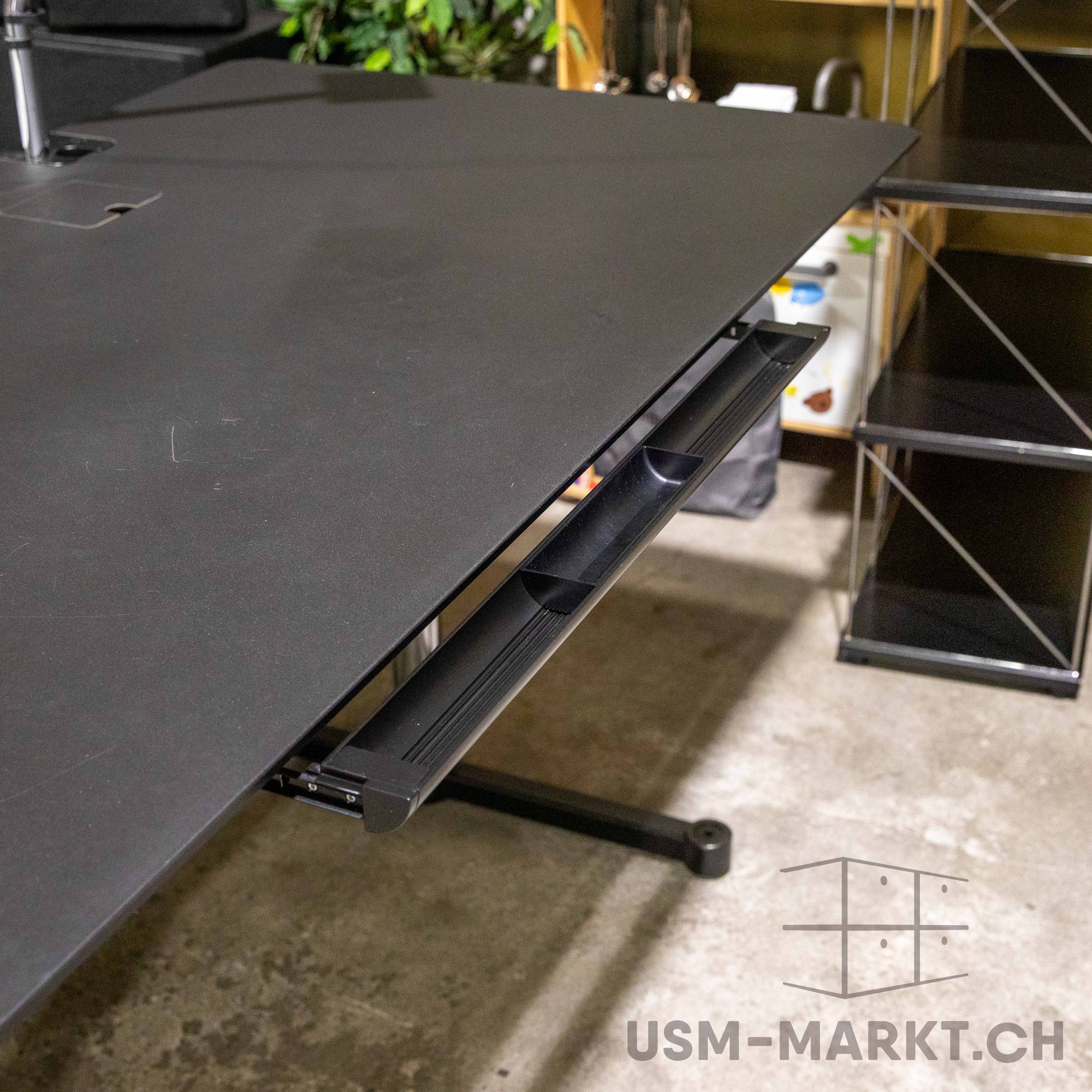 Nahaufnahme der schwarzen Linoleum-Tischplatte eines gebrauchten USM Kitos E Plus Schreibtisches mit leichten Gebrauchsspuren. Unter der Platte verläuft ein integrierter schwarzer Kabelkanal. Links oben: Öffnung für Monitorarm, Teil der Kabelklappe.