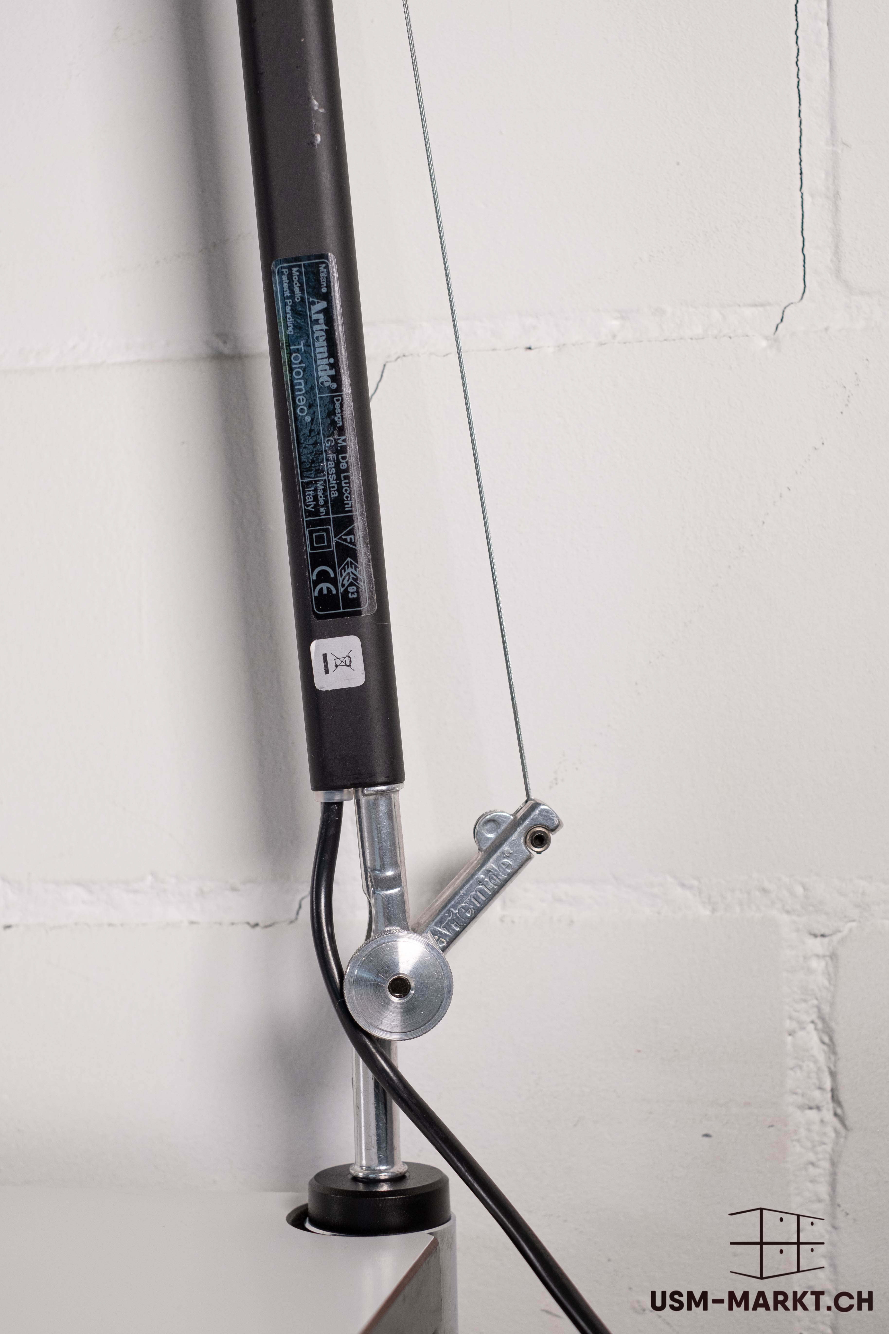 Close-up of the articulated arm of an Artemide Tolomeo desk lamp. Dark cylindrical arm with label (manufacturer, ‘Made in Italy’). Wire rope and black power cable attached to the arm at the metal joint. Light-coloured wall in the background.