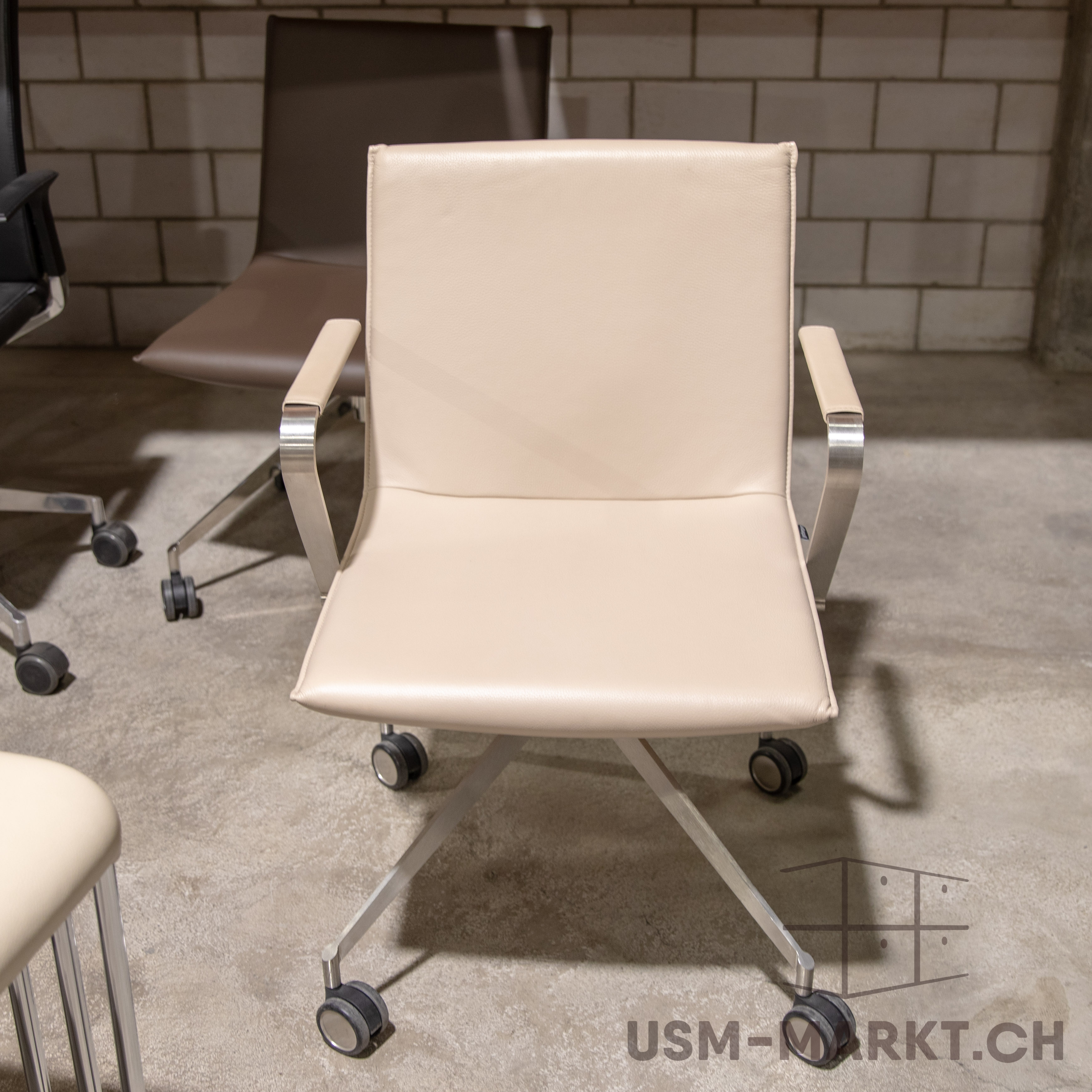 An elegant, light beige office chair with padded armrests and polished metal legs stands on a textured concrete floor. It has a five-star castor base. In the background: a light-coloured brick wall and a brown office chair.