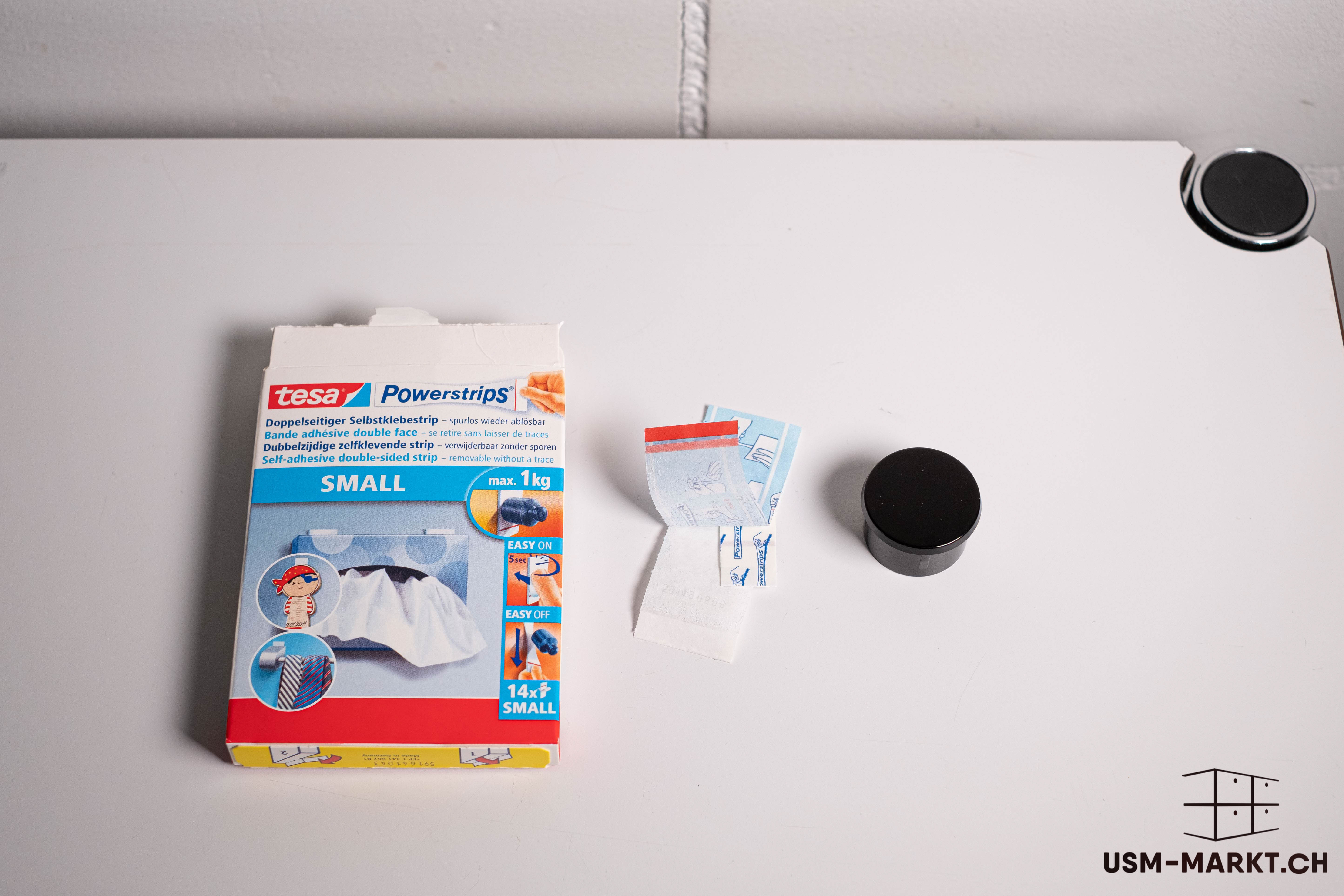 An open packet of tesa Powerstrips SMALL (double-sided adhesive strips) lies on a light-coloured surface. The red and blue packet shows examples of use. Next to it are individual Powerstrips, some of which have come loose, and a small black cylinder.