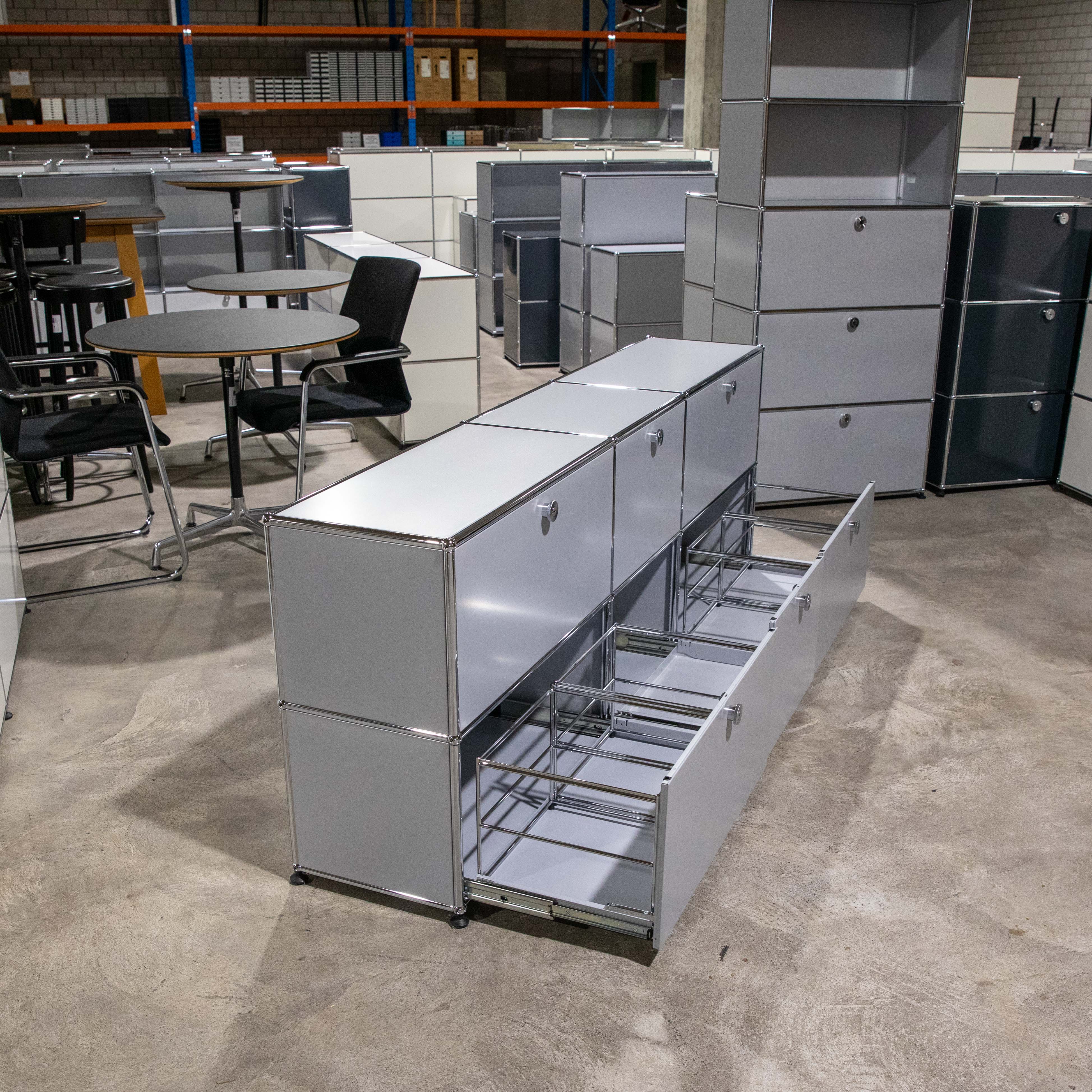 Gebrauchtes USM Haller Sideboard in Mattsilber. Drei Reihen, zwei Spalten, mehrere geschlossene Fächer sowie zwei offene Auszugschubladen mit Hängeregistratur. Im Hintergrund weitere USM Möbel im Lagerraum.