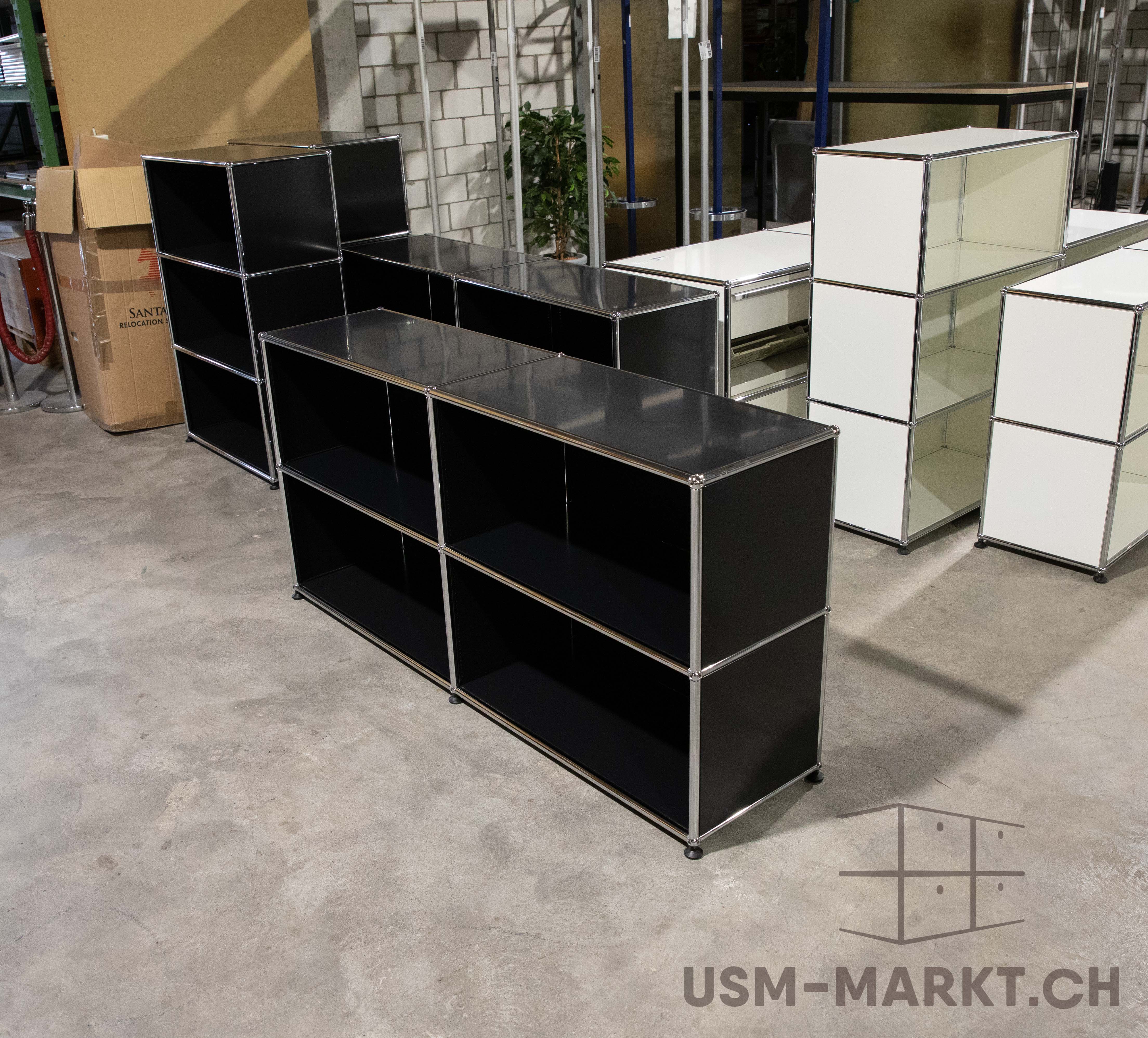 Plusieurs meubles USM Haller dans un local de stockage. Au premier plan, deux buffets noirs à compartiments ouverts. À droite, plusieurs modules USM Haller blancs de différentes tailles. Sol en béton, cartons et mur en briques à l’arrière-plan.