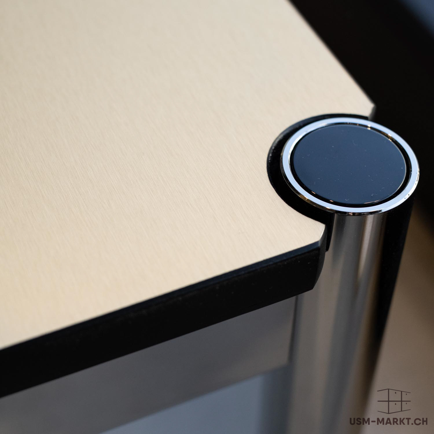 Primo piano dell'angolo di un piano tavolo chiaro beige, in impiallacciatura di legno con bordo netto e scuro. In alto a destra si vede un tappo rotondo nero lucido in un anello metallico cromato. Sotto, una gamba cilindrica.