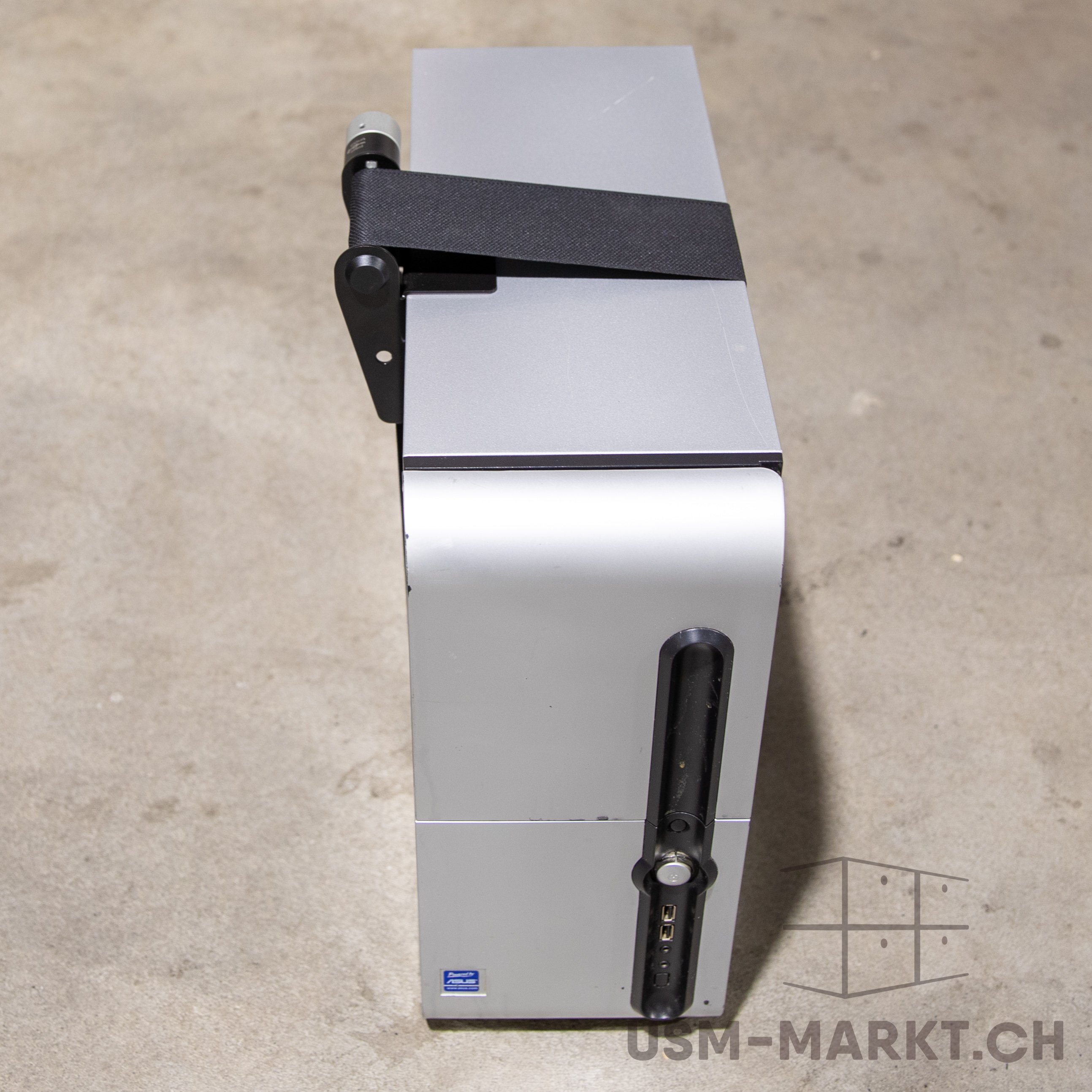 Silberner Computer-Tower auf hellgrauem Betonboden. Schwarze Transportschlaufe über die Oberseite, hinten befestigt. Vorn schwarzer Streifen mit USB- und Audiobuchsen sowie Power-Knopf. Unten links blaues ASUS-Logo.