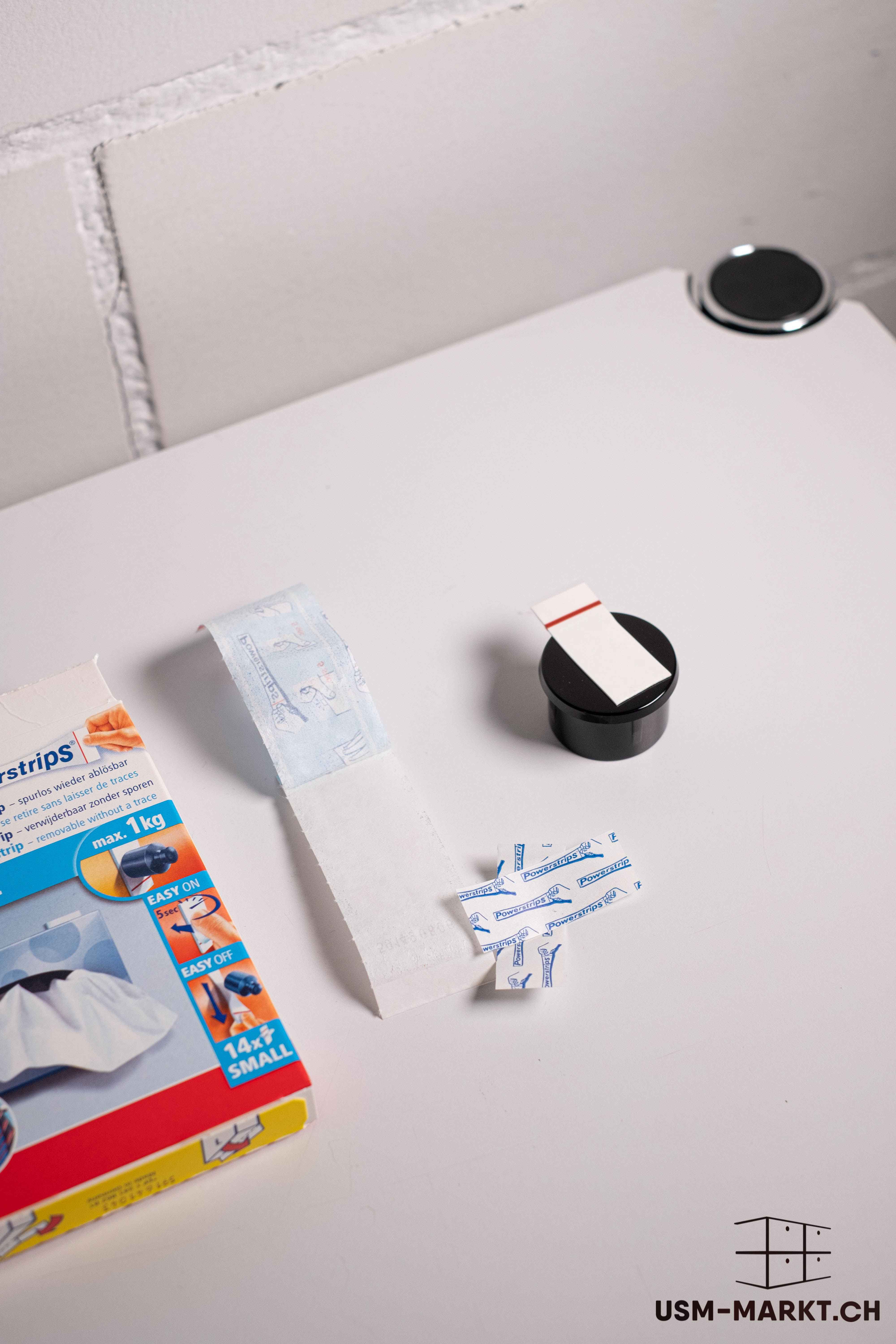 An open packet of Tesa Powerstrips and individual adhesive strips are lying on a white surface. Some of the white and blue strips have been unrolled. The red, yellow and blue packaging advertises residue-free removal and a load capacity of up to 1 kg.