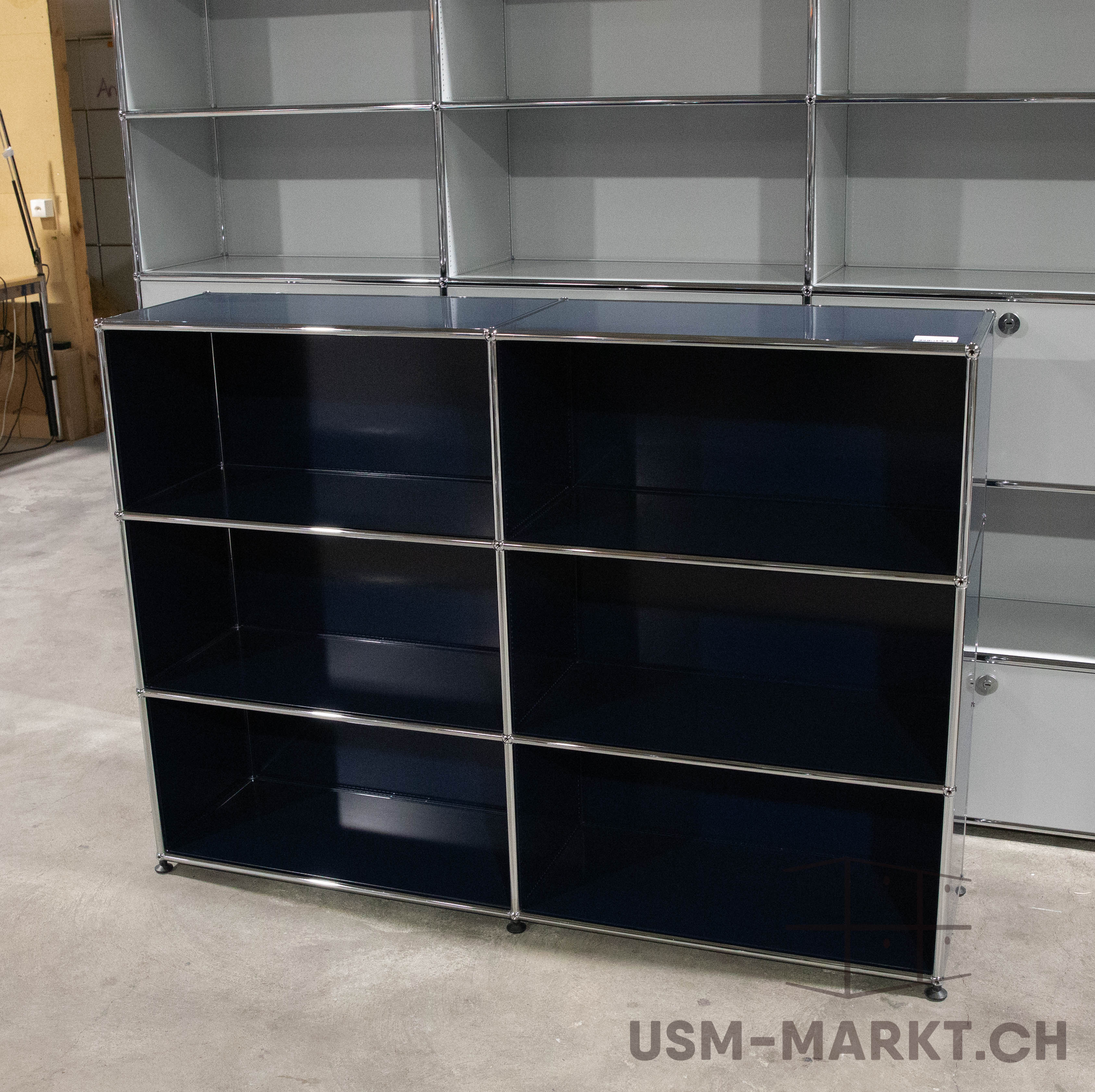 Scaffale USM Haller blu scuro con sei ripiani aperti (3 file, 2 colonne) su una base in cemento grigio. Struttura in tubi cromati lucidi, ripiani in metallo verniciato blu scuro. Sfondo: scaffale modulare grigio chiaro parzialmente visibile.
