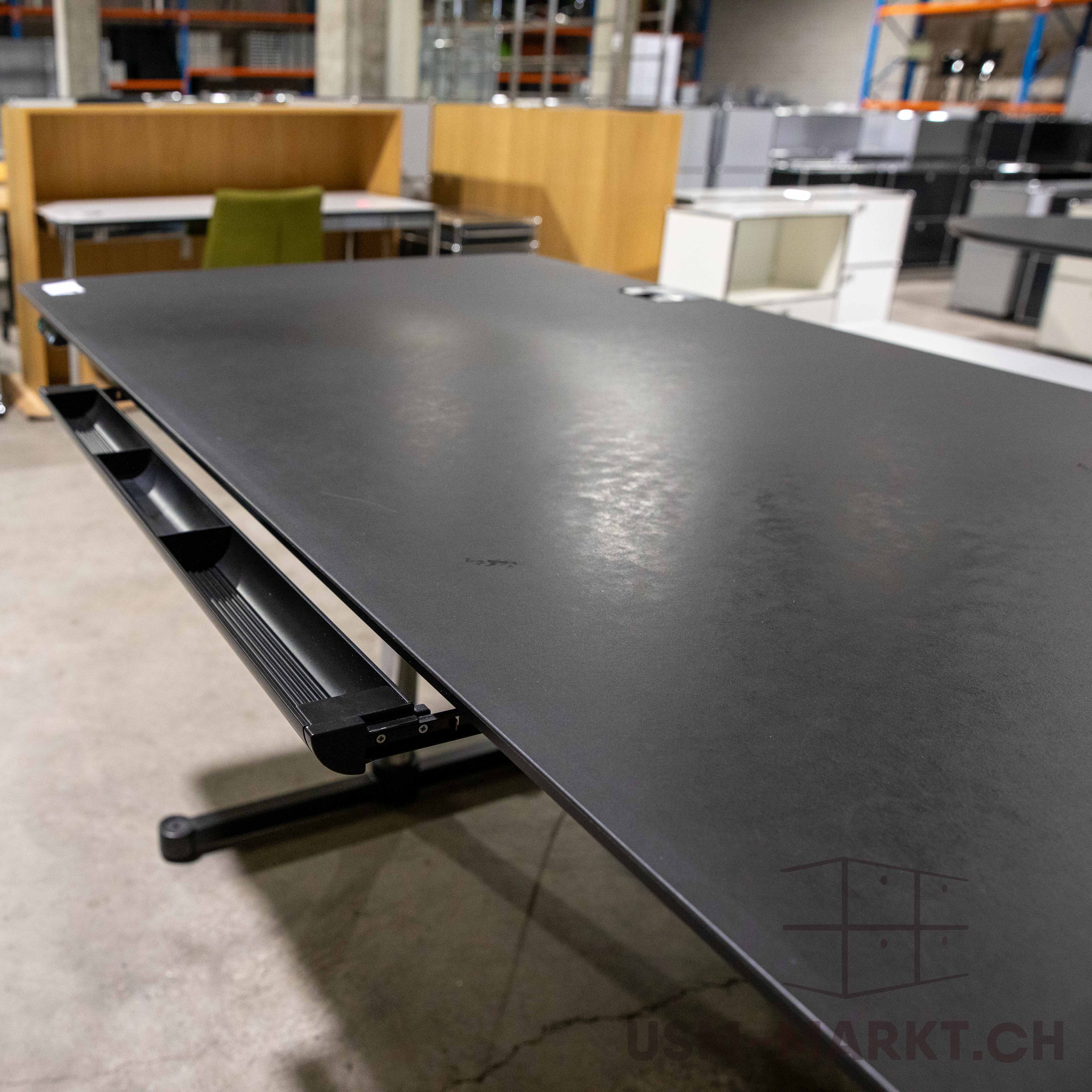 Schwarzes Linoleum-Tischblatt eines USM Kitos E Plus Schreibtisches aus leicht erhöhter Perspektive. Oberfläche mit feinen Gebrauchsspuren und Lichtreflexen. Unter dem Tischblatt ein schwarzer Kabelkanal. Hintergrund: unscharfer Lagerraum mit Möbeln.
