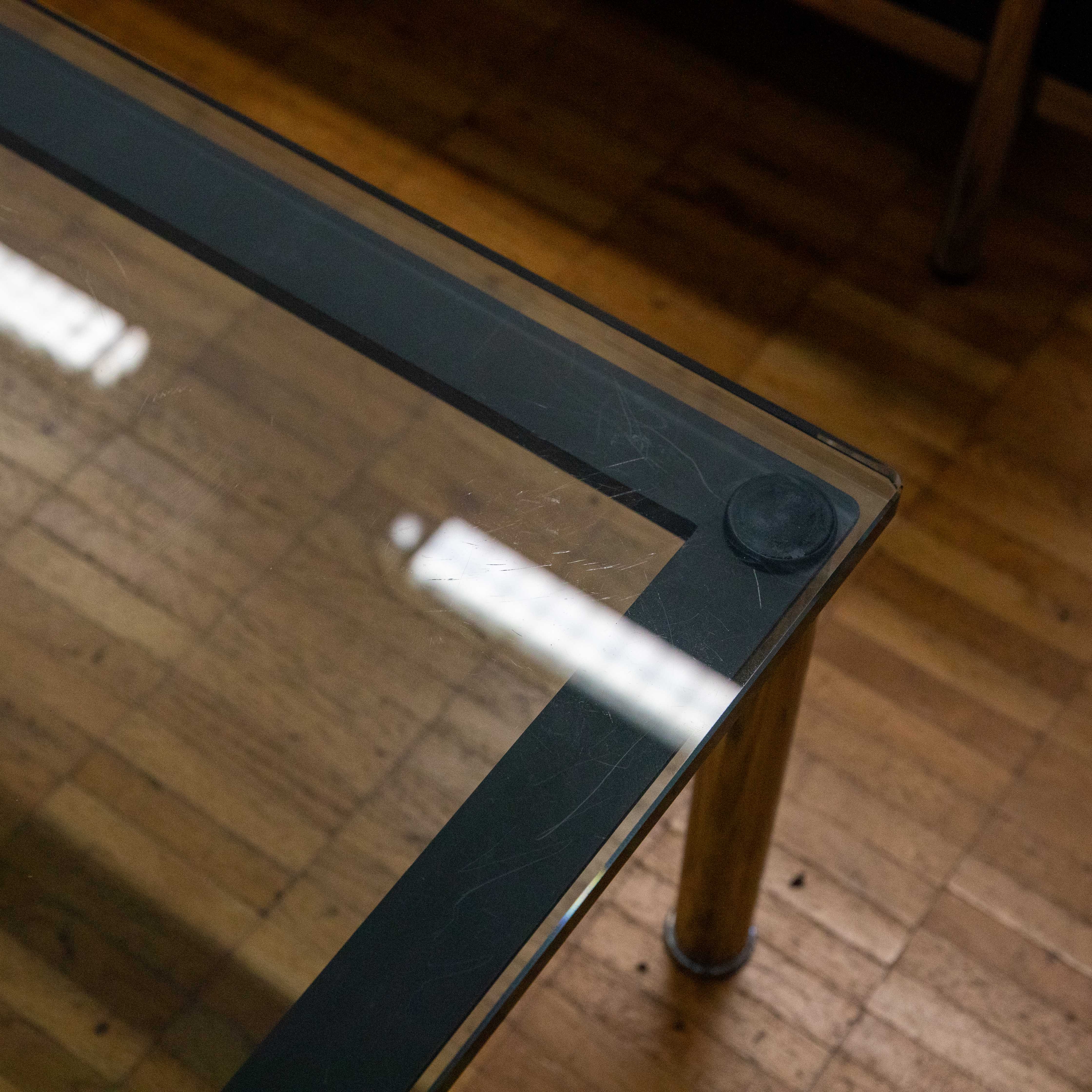 Vue détaillée d'un plateau de table en verre avec cadre métallique fin et foncé, légères rayures et reflets. À l'avant, une fixation ronde noire sur le cadre cache un pied cylindrique doré. En dessous, des planches marron clair.