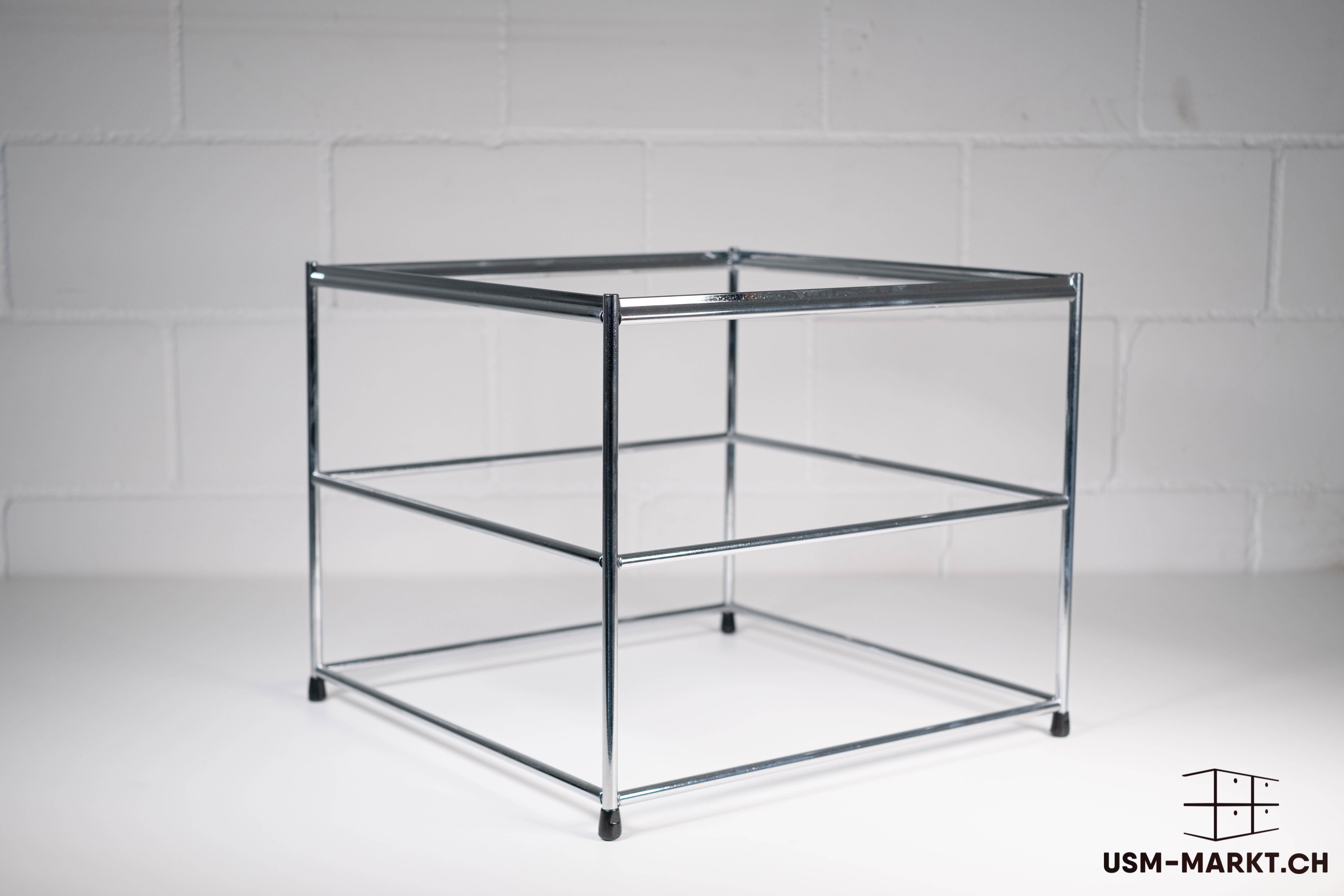 Scaffale vuoto e lucido in metallo cromato con montanti verticali e tre telai orizzontali. Il ripiano superiore è aperto, mentre i due inferiori sono telai vuoti. Piedini in plastica nera su una superficie bianca davanti a una parete di mattoni bianchi.