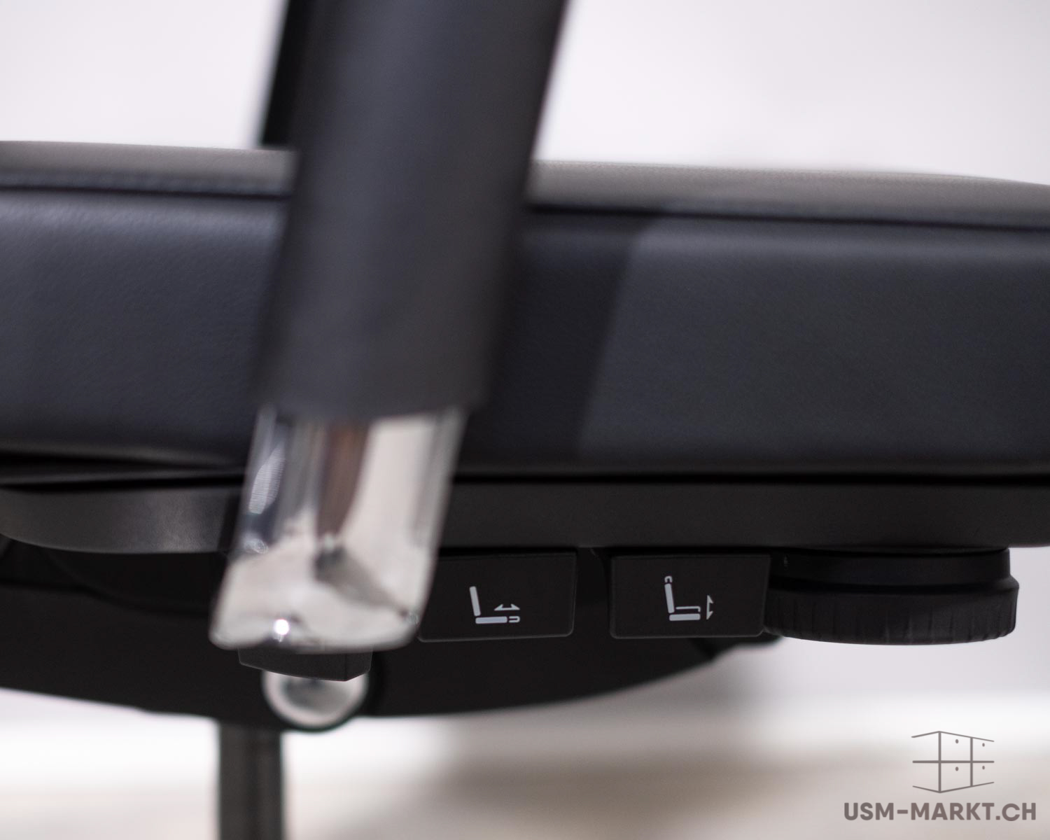 Gros plan sur la face inférieure d'une chaise de bureau noire. Deux commandes noires ornées de symboles blancs : à gauche, l'inclinaison du dossier ; à droite, le réglage de la hauteur d'assise. À côté, une molette striée et un levier chromé.