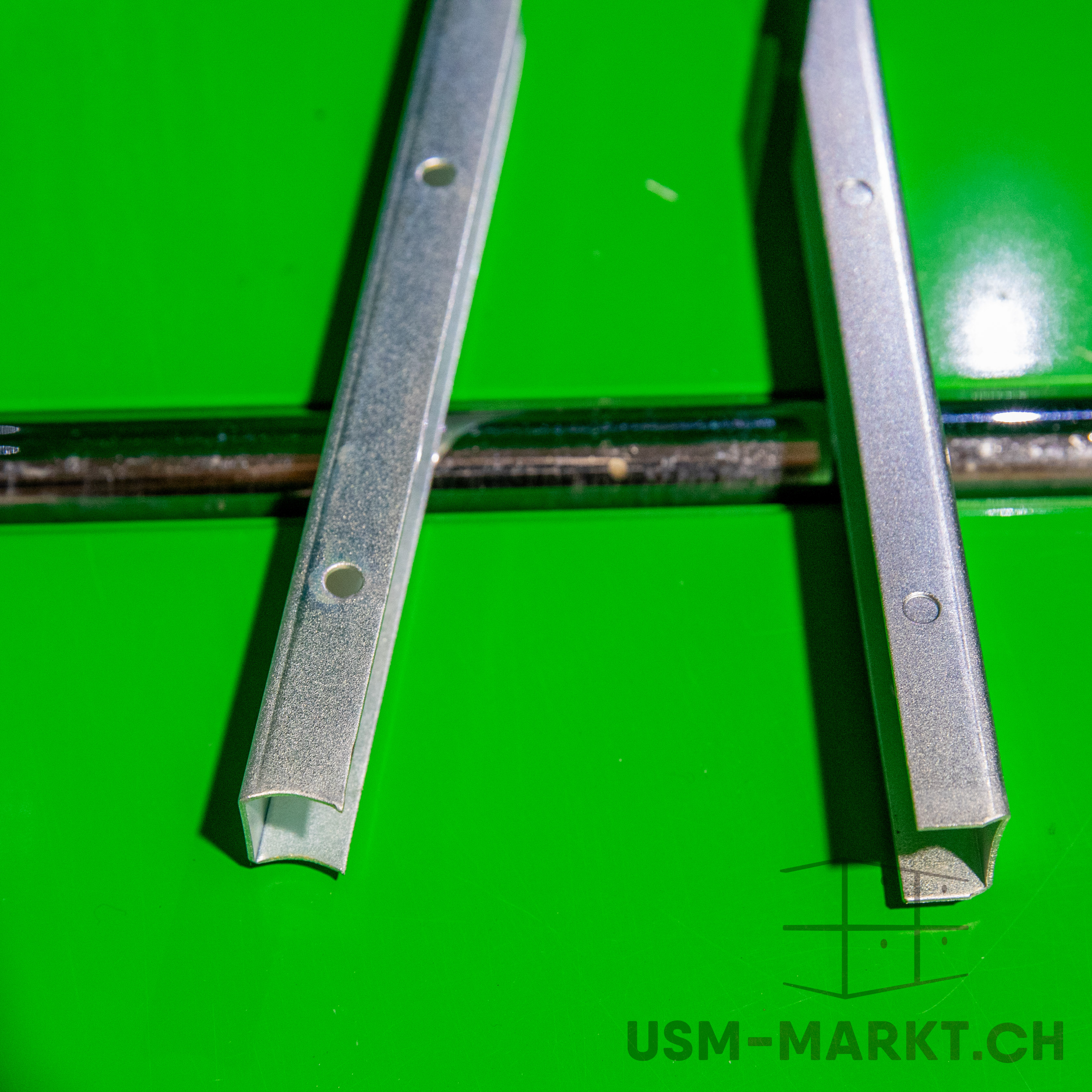 Two vertical silver metal profiles, each with a circular hole, rest on a bright green surface. A dark, shiny metal rod runs along the background. The profiles have a slightly textured, matt finish.