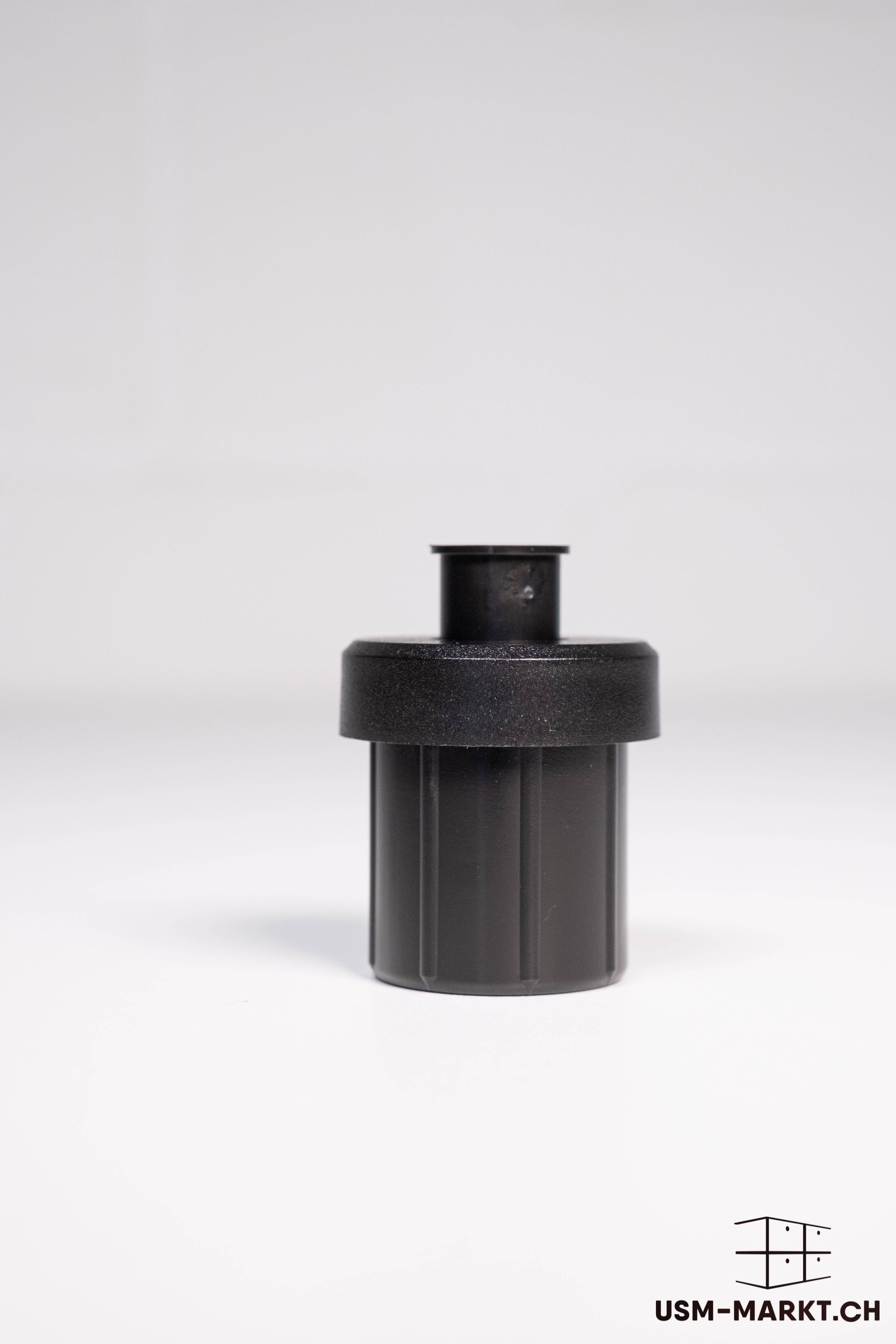 A black, cylindrical plastic part stands on a light grey background. It is ribbed at the bottom, whilst the top consists of a wider, textured disc from which a narrower, smooth cylinder protrudes vertically. Single part.
