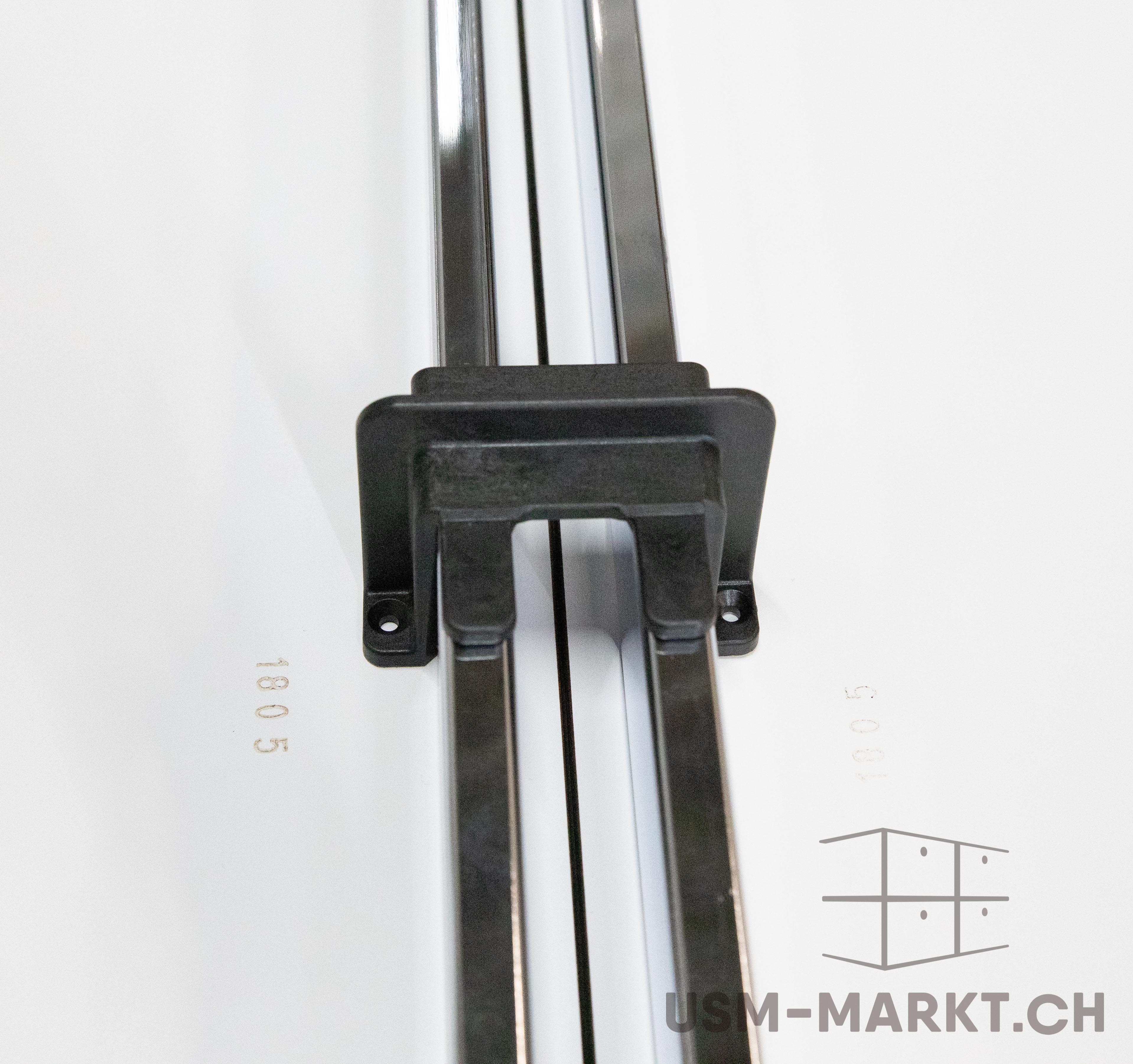 Close-up of a white surface featuring two parallel, shiny metal rods, connected by a black plastic component positioned in the centre and featuring small holes. The numbers ‘180 5’ and ‘100 5’ are faintly visible.