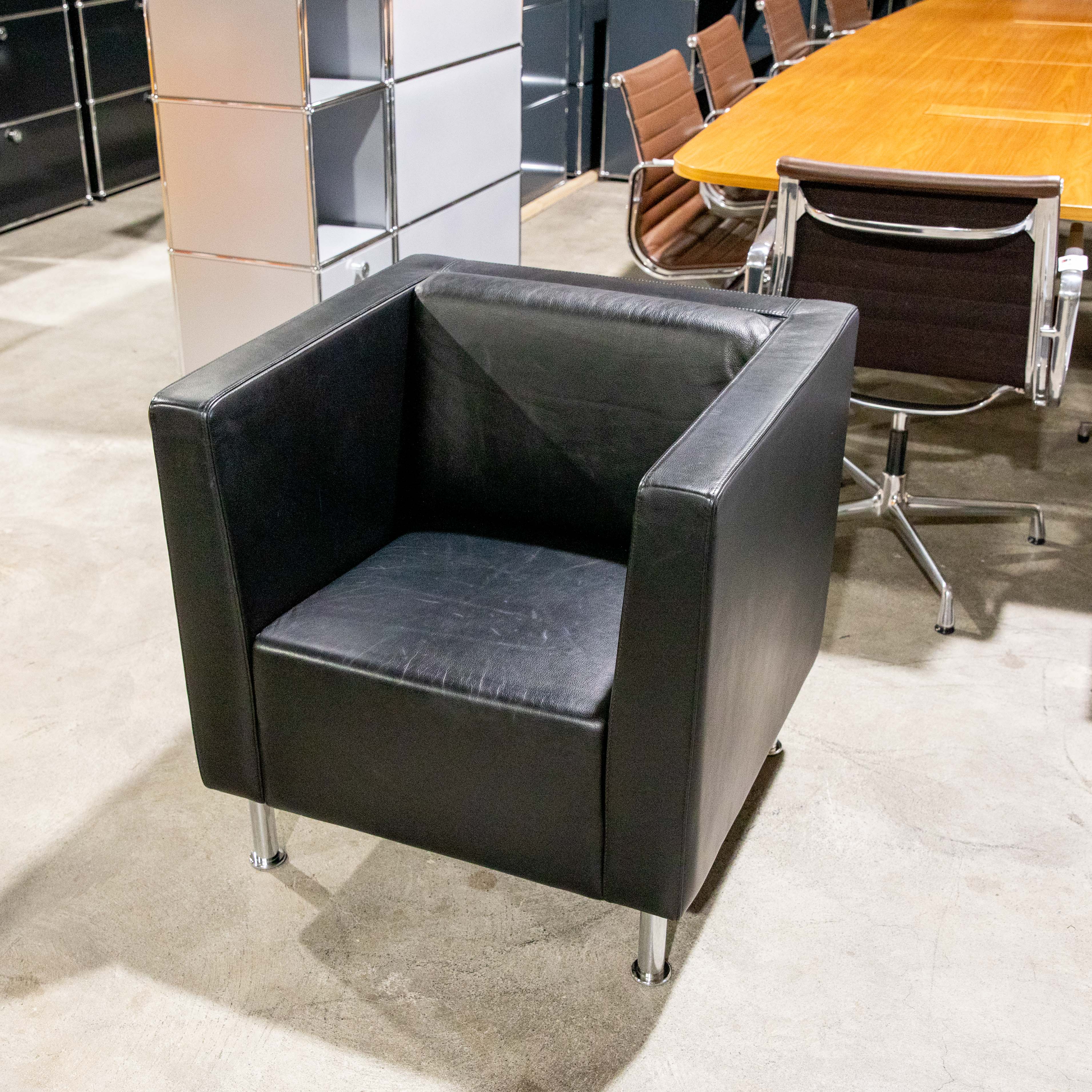 A black leather armchair with chrome legs on a light grey concrete floor. In the background, a large wooden conference table with several brown leather chairs with chrome frames to the right. To the left, modular shelving units in light grey and black.